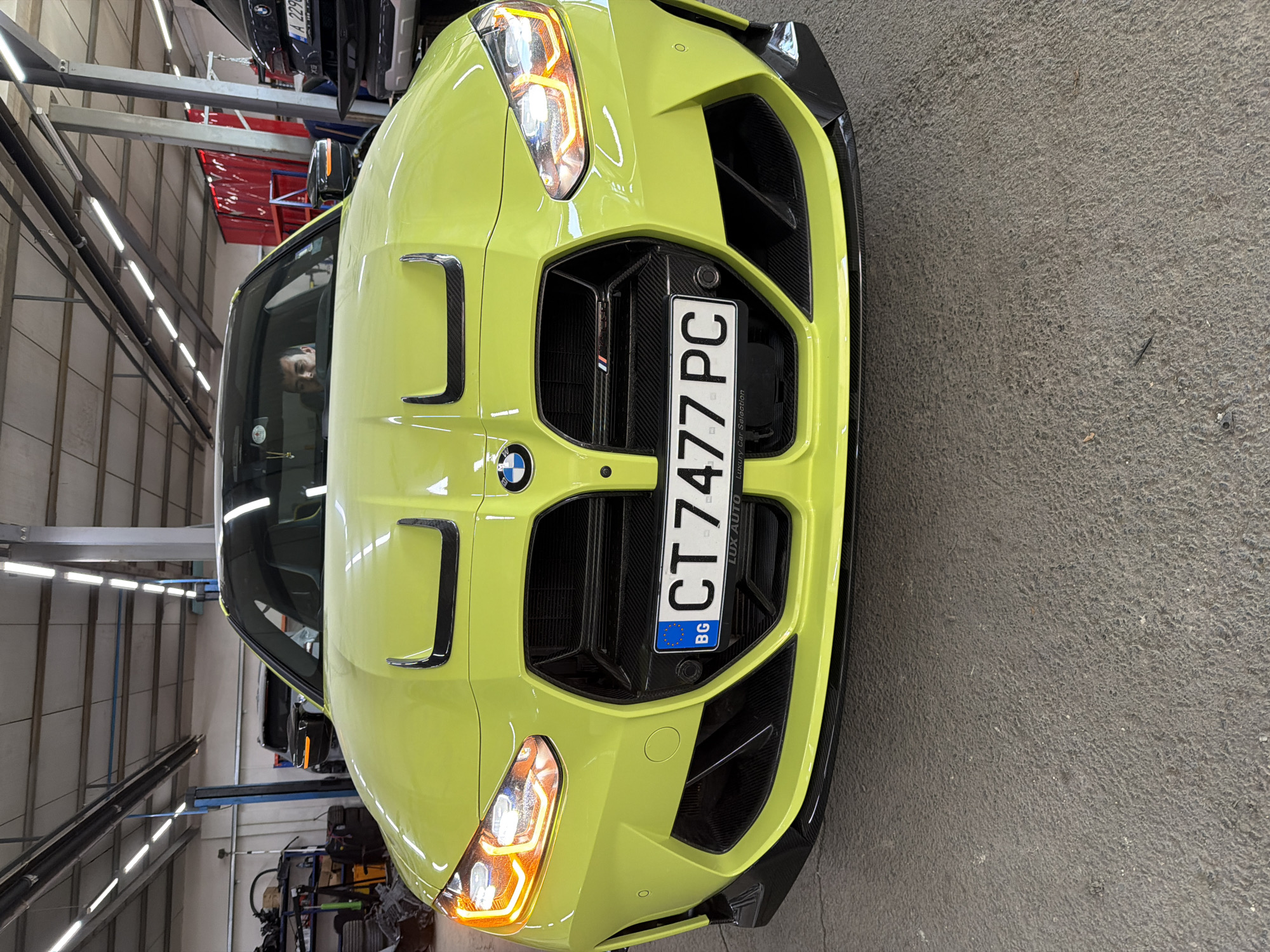 A bright, lime-green BMW M car, likely an M3 or M4, is prominently featured, taking up the majority of the frame. The car is seen from the front, with its large, vertical kidney grilles and the BMW logo clearly visible. Carbon fiber accents adorn the grilles, hood vents, and lower bumper area. Its headlights are illuminated with an amber hue.
A European license plate is mounted centrally on the front grille, displaying "BG" for Bulgaria, and the registration number "CT 7477 PC". Below the registration, the plate frame includes text "Lux Auto" and "Luxury Car Selection".
The car is parked indoors, in what appears to be an automotive workshop, garage, or showroom. The environment is well-lit by numerous overhead fluorescent or LED strip lights, which reflect brightly on the car's glossy surface. In the background, metal shelving and other vehicles are visible. One car is suspended on a lift, with its rear bumper and a partial license plate ("A 2296 B") discernible.
A person's face is partially visible inside the lime-green car, reflected in the windshield or side window, looking forward. The overall scene suggests a professional setting for vehicle maintenance, display, or preparation.
