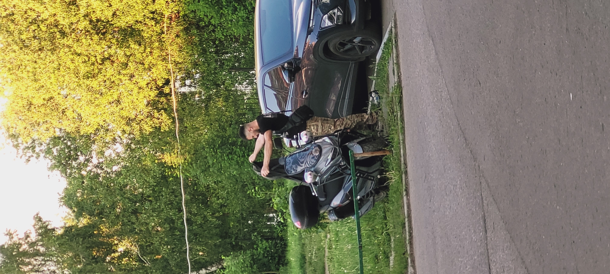 The image depicts an outdoor daytime scene in the city of Unknown, Unknown, featuring a man standing next to a maxi-scooter with a car parked nearby, set against a backdrop of dense greenery and an asphalt road.
In the center of the frame, an adult male with short dark hair and facial stubble is visible. He wears a black short-sleeved t-shirt, camouflage-patterned trousers, and a black cross-body shoulder bag. His arms are raised, and he is holding a black garment or item, possibly a jacket or bag, with both hands, appearing to be either putting it on or taking it off.
To the man's left is a gray maxi-scooter with a large windscreen, prominent headlights, chrome-accented mirrors, and a black hard-shell top case mounted at the rear. The scooter is parked on a grassy verge, leaning slightly, with its lower front section partially obscured by a horizontal green metal bar in the foreground and a wooden stake behind it.
To the man's right, a dark-colored SUV (possibly black or dark blue) is parked, with its front and side partially visible. Reflections of the surrounding trees are apparent on its windows. On the grass near the car's front tire, a small blue and white object is discernible.
The background consists of a dense wall of various green trees, predominantly deciduous, with sunlight illuminating some of the foliage, suggesting a sunny day. A thin, light-colored cable or wire stretches horizontally across the upper mid-ground. The foreground shows a strip of unkempt green grass containing some dandelions, bordering an asphalt road that occupies the lower quarter of the image. The road surface is gray with visible cracks and faint lines.