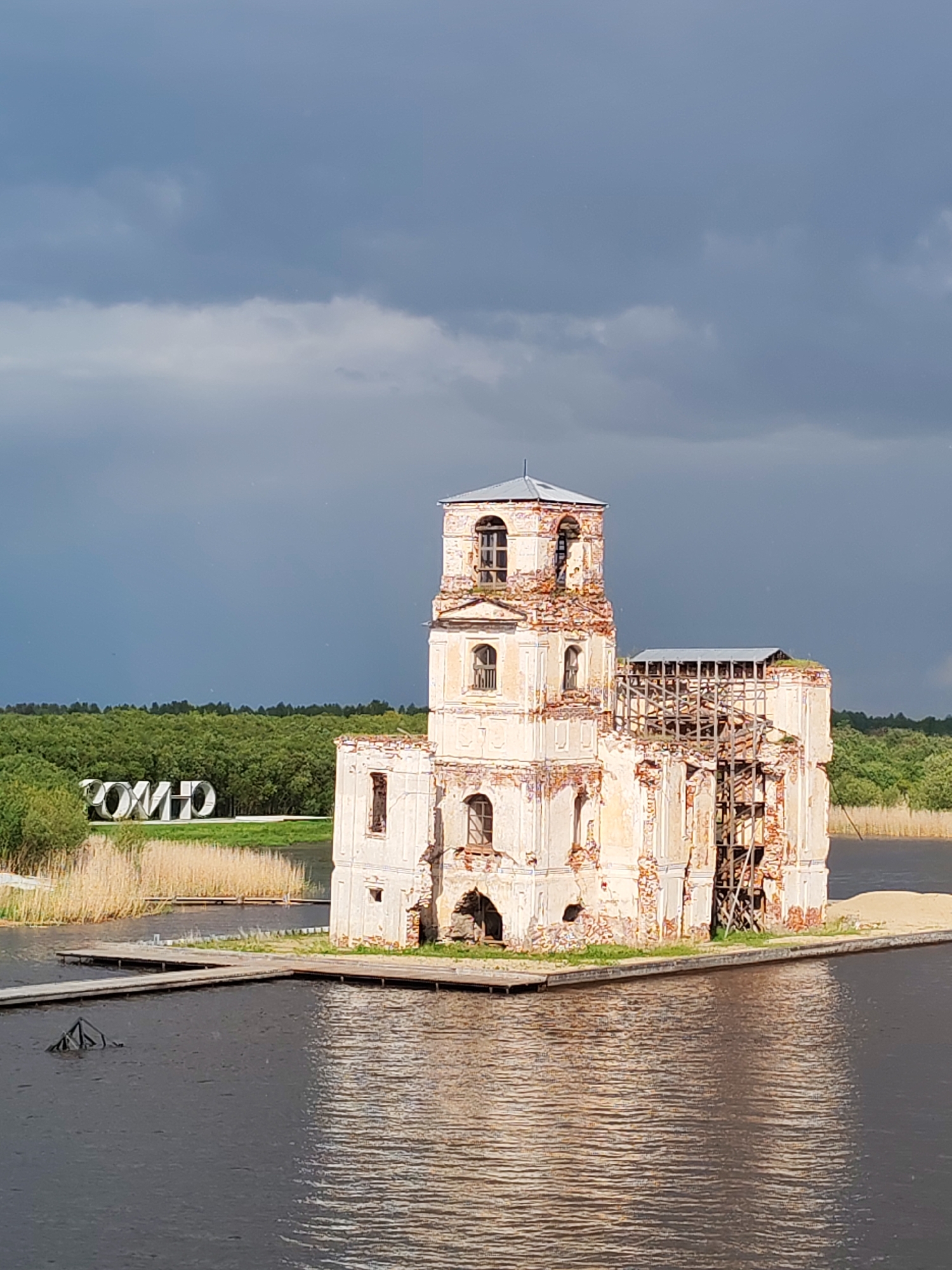 The image depicts the partially submerged and dilapidated ruins of the Church of the Nativity in Krokhino, Russia. The structure, composed primarily of white and red brick, stands on a small plot of land surrounded by water, accessible by wooden walkways. The church's tower and parts of its main structure are significantly deteriorated, with visible decay and crumbling sections. A framework of scaffolding and a makeshift roof cover a portion of the ruins, suggesting ongoing or planned preservation efforts. The foreground features the dark, rippled surface of the water, while the background includes a grassy area with reeds, trees, and an art installation in the distance reading “КОММО”, possibly in Cyrillic. The sky above is overcast, with dark, ominous clouds indicating an approaching storm.