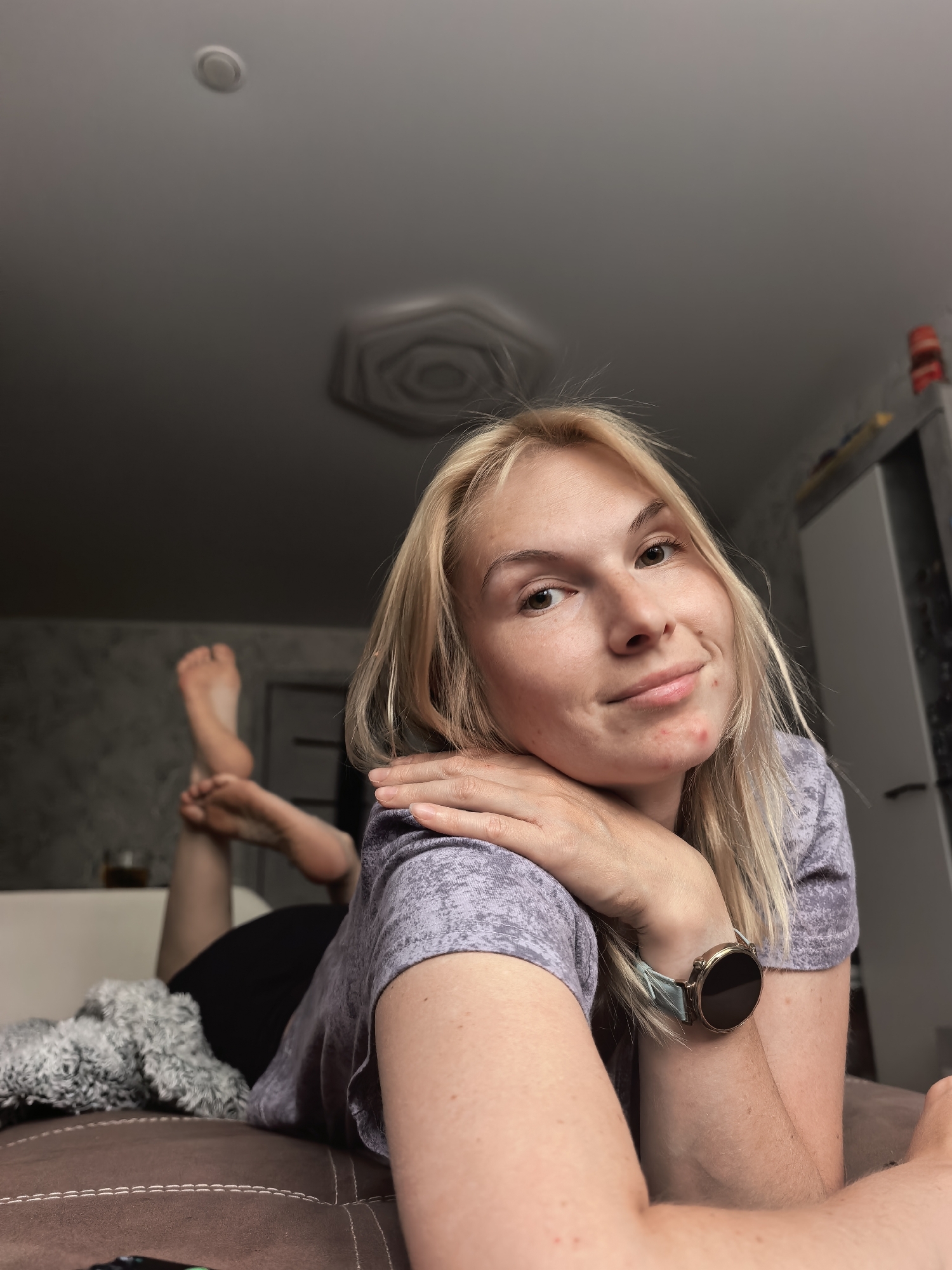 A young blonde woman is lying on her stomach on a brown couch in an indoor setting. Her head is facing the camera, and her hands are crossed under her chin. She is wearing a light purple t-shirt and a dark-colored bottom. A smartwatch is visible on her left wrist. Her feet are raised and crossed in the background. A grey blanket is next to her on the couch. A glass is next to the feet. In the background, there's a blurry view of a room, a light fixture on the ceiling, and a white cabinet. The location of the image is unknown.