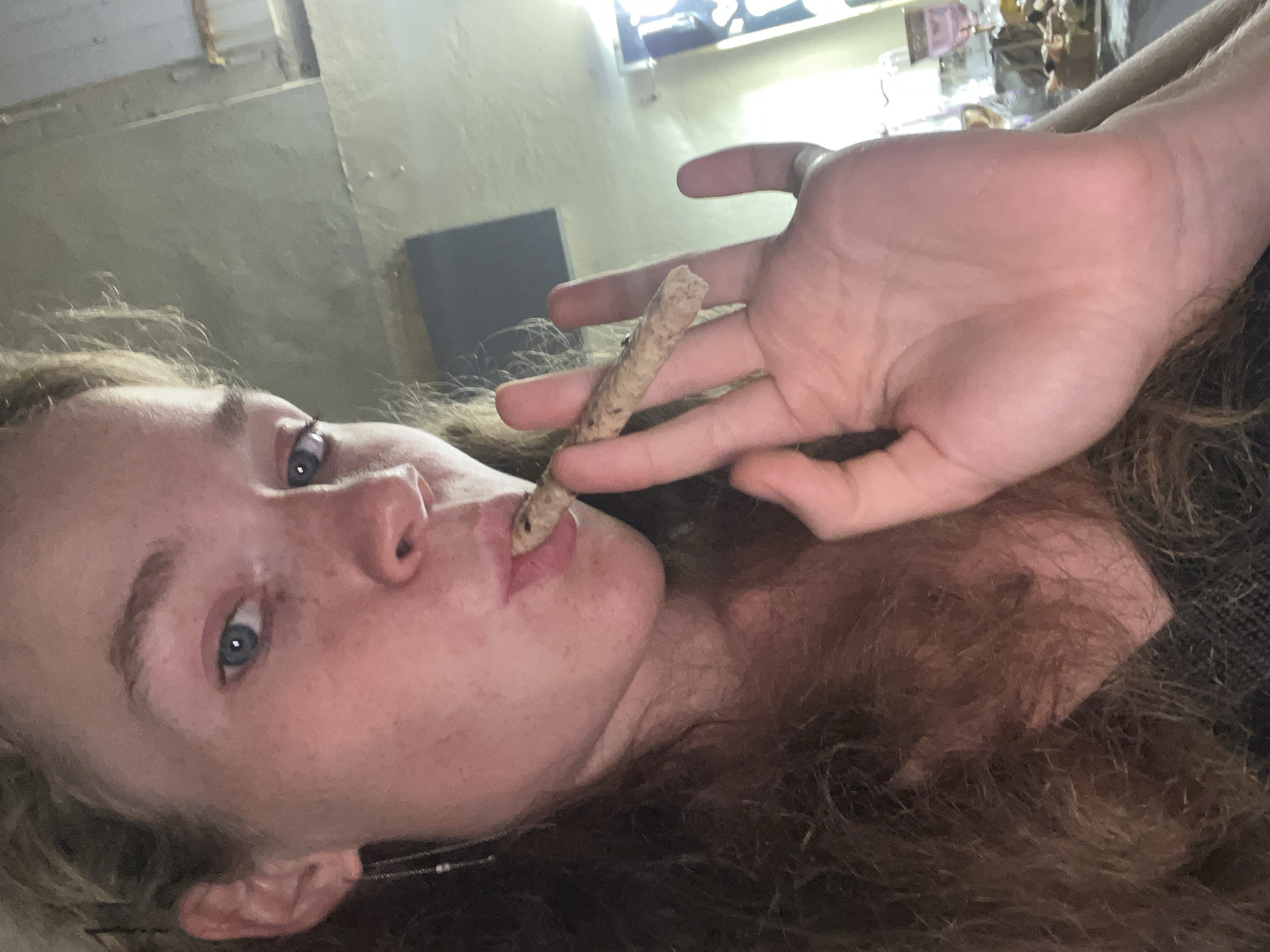 The media file shows a close-up, overhead shot of a person with long, reddish-brown hair. The person is lying down, with their face tilted towards the camera. They have light skin with freckles and blue eyes. Their hand is raised to their mouth, holding a rolled item, possibly a cigarette or joint, to their lips. The person's expression is difficult to discern due to the angle and focus.
The background appears to be an indoor setting, with rough, textured walls and a bright light source at the top of the frame, suggesting daylight. There are hints of objects in the background, possibly shelves or decorative items, but they are out of focus and indistinct. The overall lighting is dim except for the bright area at the top.
The scene suggests a moment of personal activity or relaxation. The focus is on the person's face and hand, drawing attention to the action of holding the rolled item. There is no visible text in the image. The context provided, Budva, Montenegro, does not offer specific visual cues within the image itself.