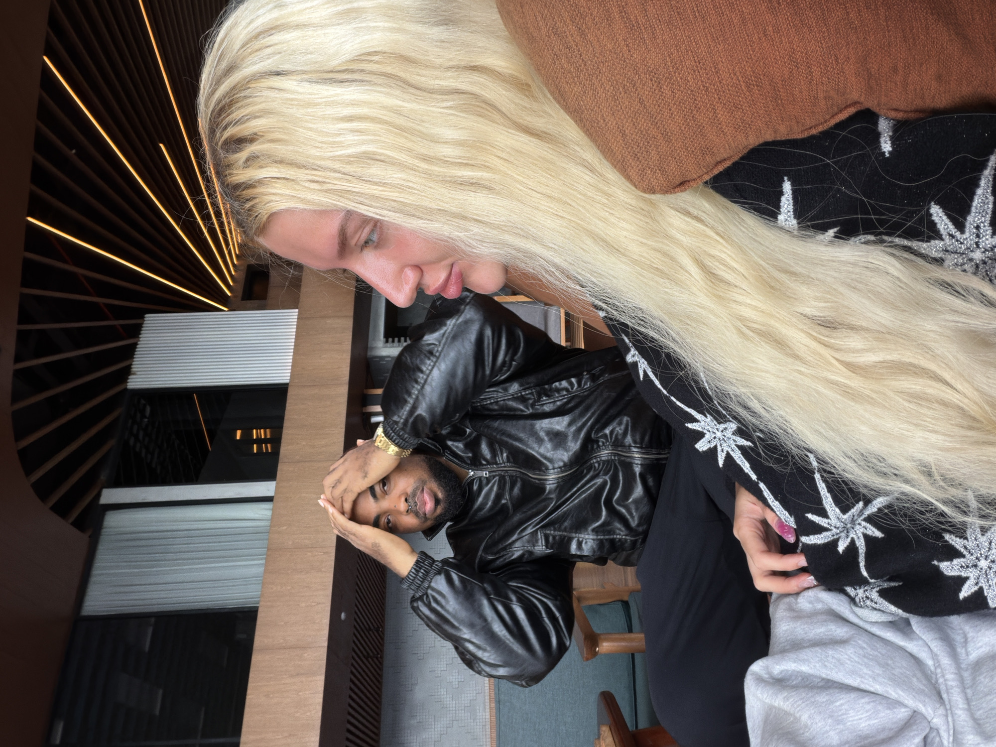 An overhead shot captures a man in a black leather jacket with his hands on his head, sitting in a chair. To his right, a woman with long blonde hair is in profile, looking away from the camera. The background features a modern interior with wooden paneling and ceiling lights. The setting appears to be a cafe or restaurant in Kintamani, Indonesia, indicated by the distinct architectural style and ambient lighting often found in establishments catering to tourists in the region.