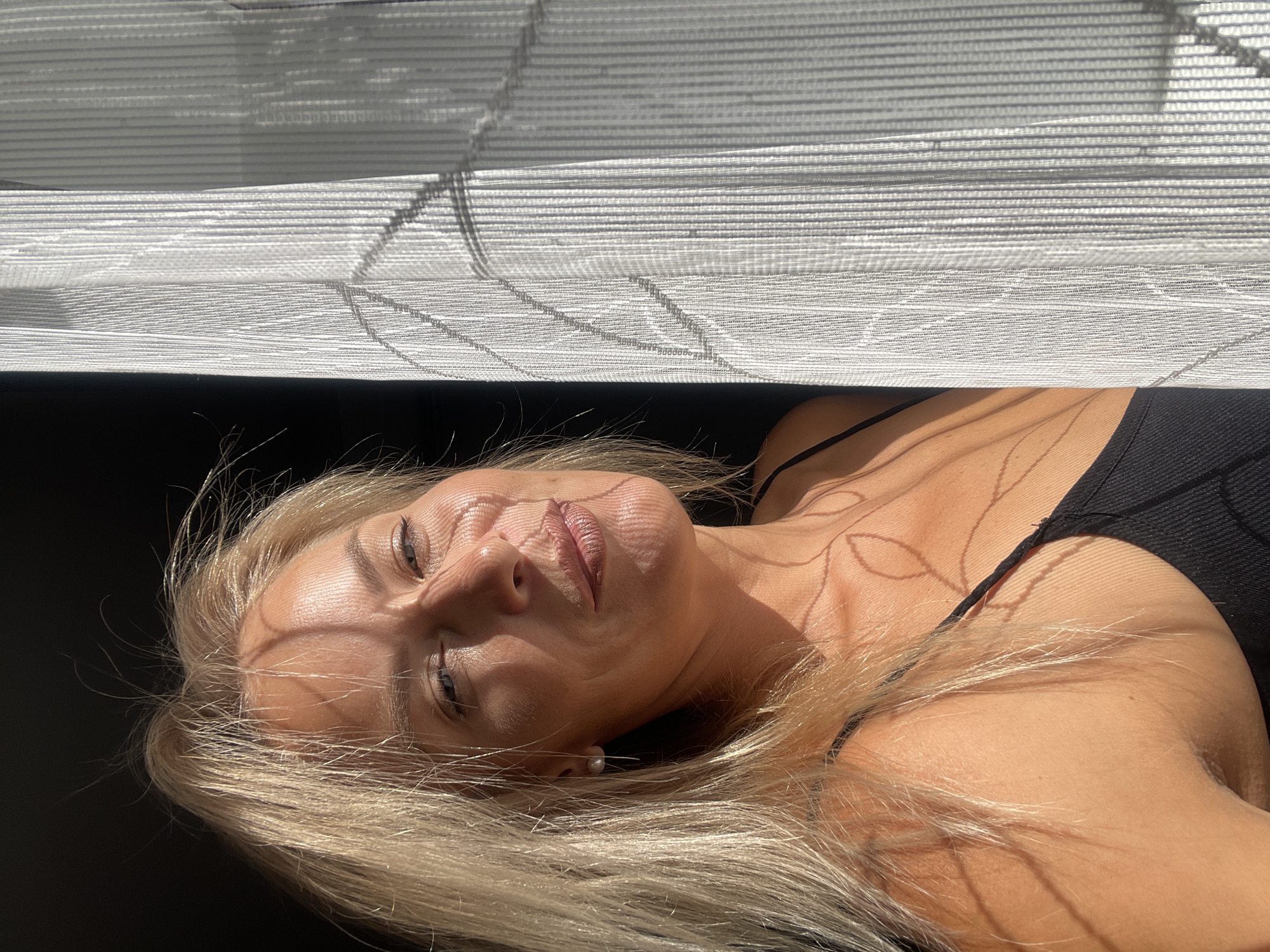 A middle-aged Caucasian woman with blonde hair and tanned skin poses for a photo. She is wearing a black tank top and a pearl earring. The woman is positioned to the left of the frame, with her face illuminated by sunlight casting shadows from a sheer curtain. The background is split between a dark area on the left and the white curtain on the right. The image appears to be taken indoors, possibly in Krasnodar, Russia, during daylight.