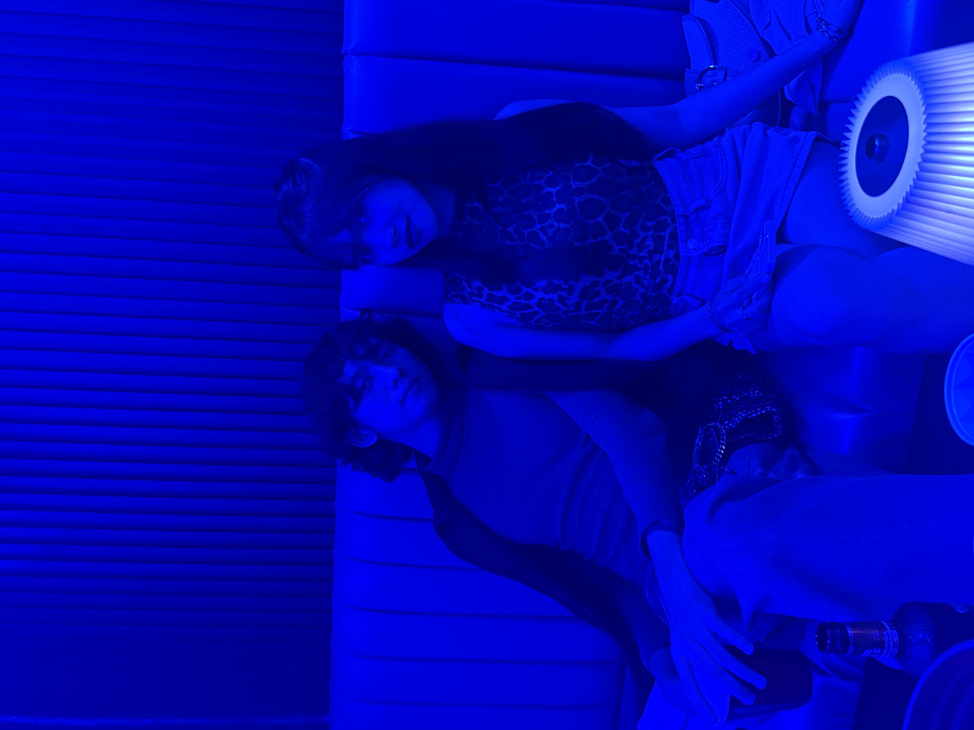 Two individuals, a male and a female, are seated on a blue couch in a dimly lit room illuminated by blue lighting. The male, on the left, has dark, curly hair and is wearing a dark jacket over a lighter-colored top and jeans. He is holding a red cup in his left hand. The female, on the right, has long dark hair and is wearing a leopard-print sleeveless top and a denim skirt. She is looking towards the camera. In front of the couch, a bottle and a white lamp with a ribbed shade are visible. The background features a textured blue wall. The environment suggests an indoor setting, possibly a lounge or a bar. Based on the provided information, this scene is located in Davao City, Philippines.