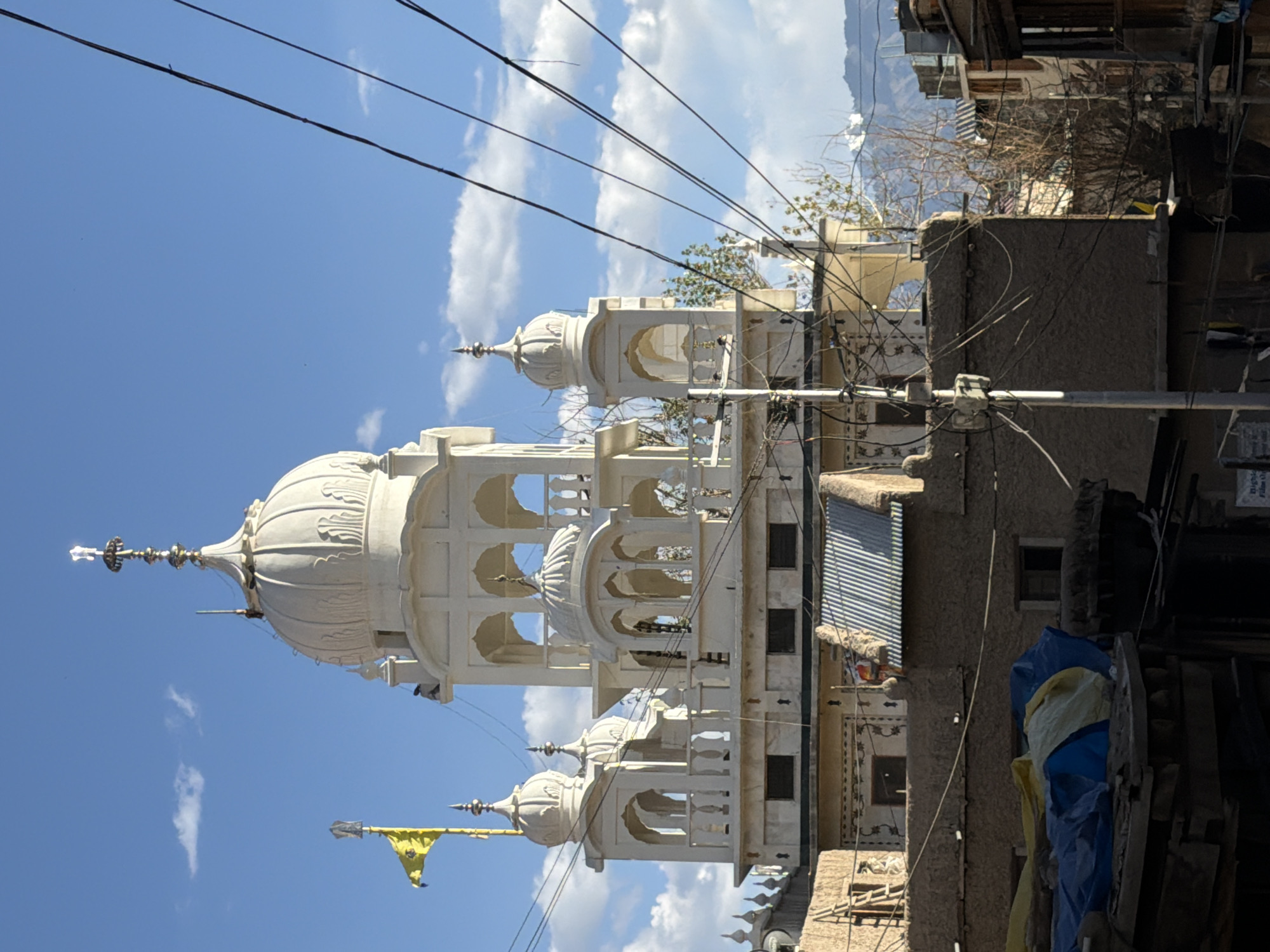 The image shows a white Sikh gurdwara in Leh, India, with a yellow Sikh flag flying from its roof. The gurdwara is elaborately decorated with domes and arches, and is surrounded by other buildings, some of which appear to be made of mud brick. Power lines are visible in the foreground, crisscrossing the frame. The sky is blue with scattered clouds, and mountains can be seen in the background. The sun is shining, casting shadows on the buildings. There are no people clearly visible in the image. The overall scene depicts a religious building in a somewhat rustic urban environment.