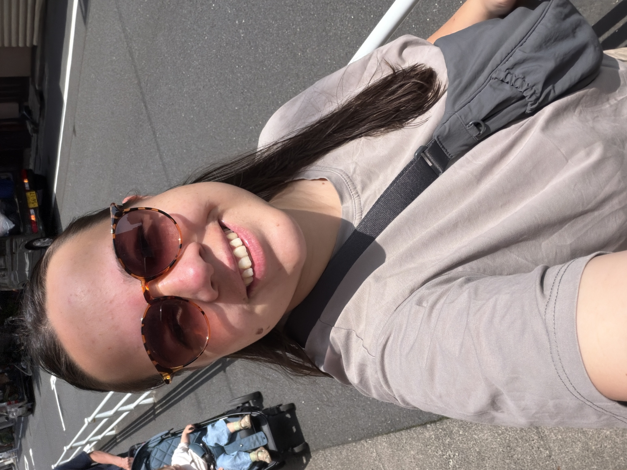 A selfie taken outdoors in Chuo, Japan, shows a woman wearing sunglasses and a light brown t-shirt with a gray crossbody bag slung over her shoulder. She is smiling at the camera. To her left, a child sits in a stroller, dressed in blue pants and a white shirt. The background features a paved road with a white line, a sidewalk, a white railing, and vehicles parked along the street. The lighting suggests it is daytime.