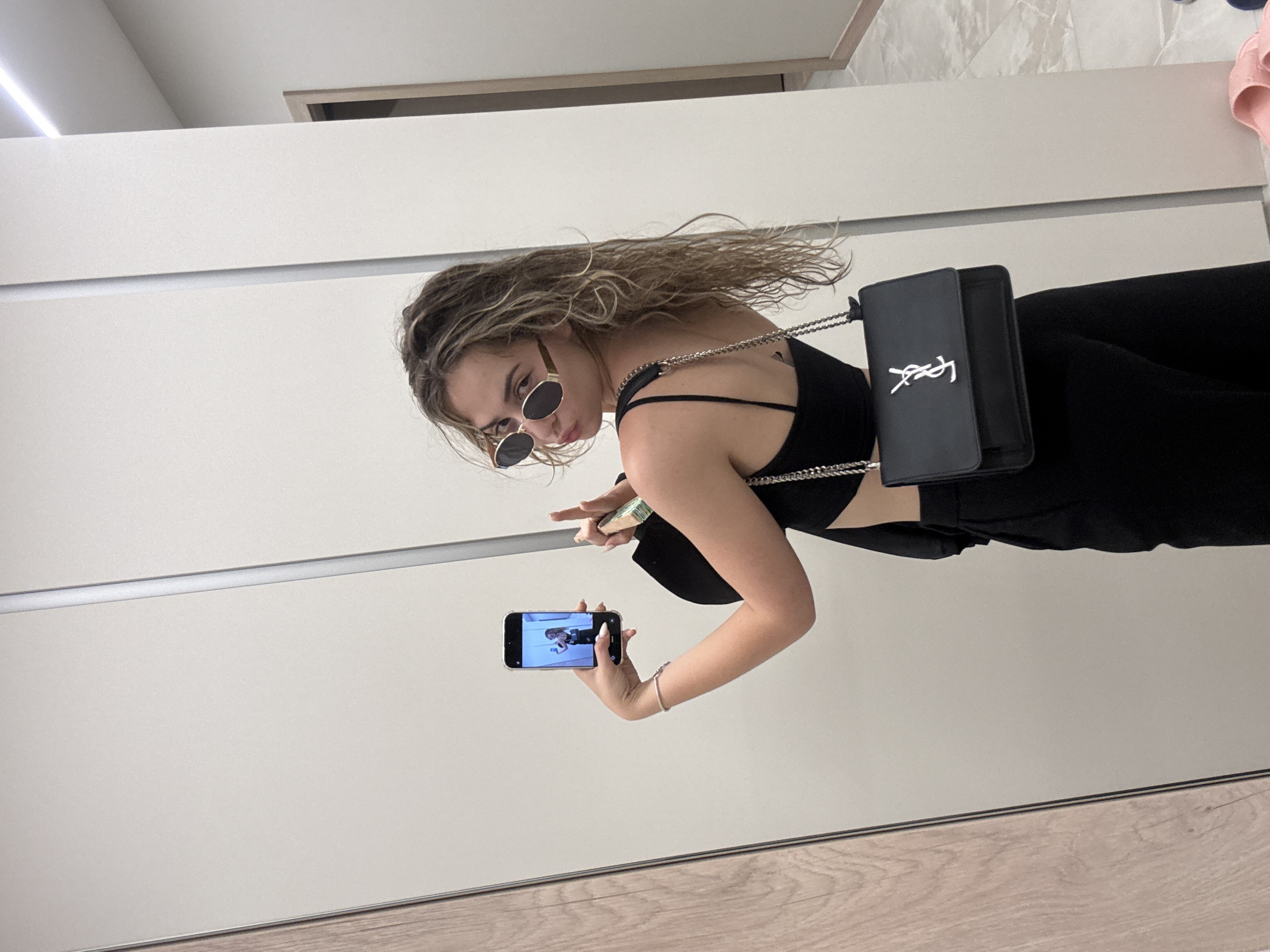 A young woman is captured in a mirror selfie within an interior setting, identified as the city of Sofia, Bulgaria. She stands with her back partially turned, looking over her right shoulder towards the camera with a slight pout. Her medium-length, wavy, light brown hair cascades down her back. She wears oval-framed sunglasses, a black spaghetti-strap crop top revealing her midriff, and black wide-leg or straight-leg pants. A black shoulder bag with a silver chain strap and a prominent silver "YSL" logo hangs from her left shoulder. On her left wrist, a silver-colored bracelet is visible. She holds a black smartphone in her left hand, which displays her own reflection. In her right hand, she holds an elongated, light-colored object, possibly a perfume bottle or hand sanitizer. The background is dominated by the mirror reflecting a light-colored wall or wardrobe doors with a vertical seam. To the far left, a strip of light-colored wood paneling is reflected. To the far right, a doorway with a light-colored frame and a dark interior is visible in the reflection, along with a linear ceiling light fixture. The floor is covered with light-colored, subtly patterned tiles, and a pair of pink sandals are visible on the floor to the right.
