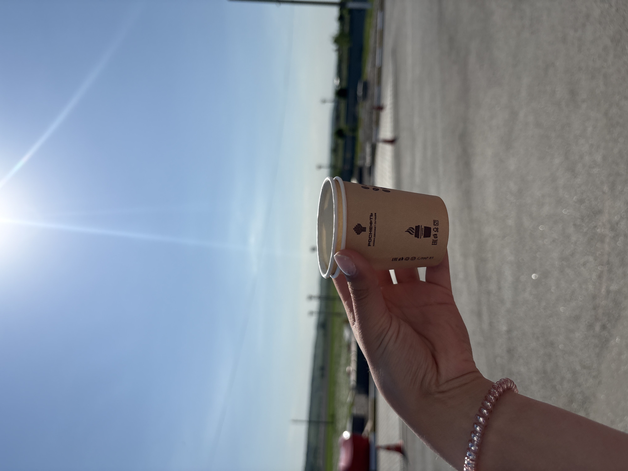 A person holds a brown paper coffee cup in their right hand against a bright blue sky. The cup features printed branding in Cyrillic script along with an image of a steaming coffee cup. A pink beaded bracelet adorns the person's wrist. The background shows an out-of-focus asphalt parking lot with distant utility poles and green vegetation. The bright sun, with its associated lens flare, dominates the left side of the frame. The location is an outdoor area possibly in Mar'evka, Russia, based on the branding language on the cup.