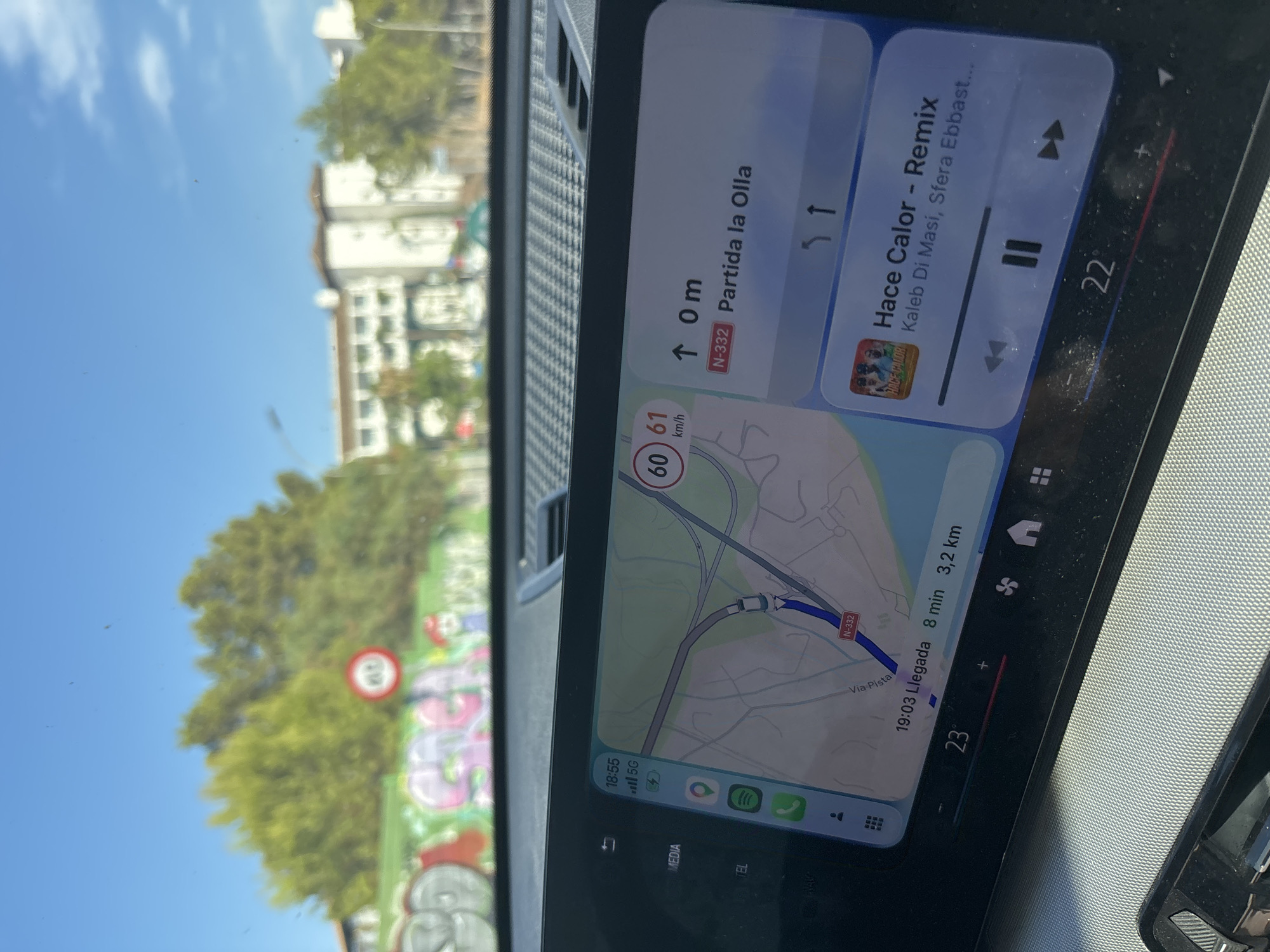 The image shows a car's dashboard display with navigation and music applications open. The car is traveling on a road with graffiti on a wall to the left. In the background, there are trees and buildings. The navigation shows a route on the N-332 road towards "Partida la Olla" in Altea, Spain, with an estimated arrival time of 19:03 and a distance of 3.2 km. The current speed is displayed as 60-61 km/h. The music player shows the song "Hace Calor - Remix" by Kaleb Di Masi, Sfera Ebbast... The ambient temperature inside the car is 23 degrees Celsius.