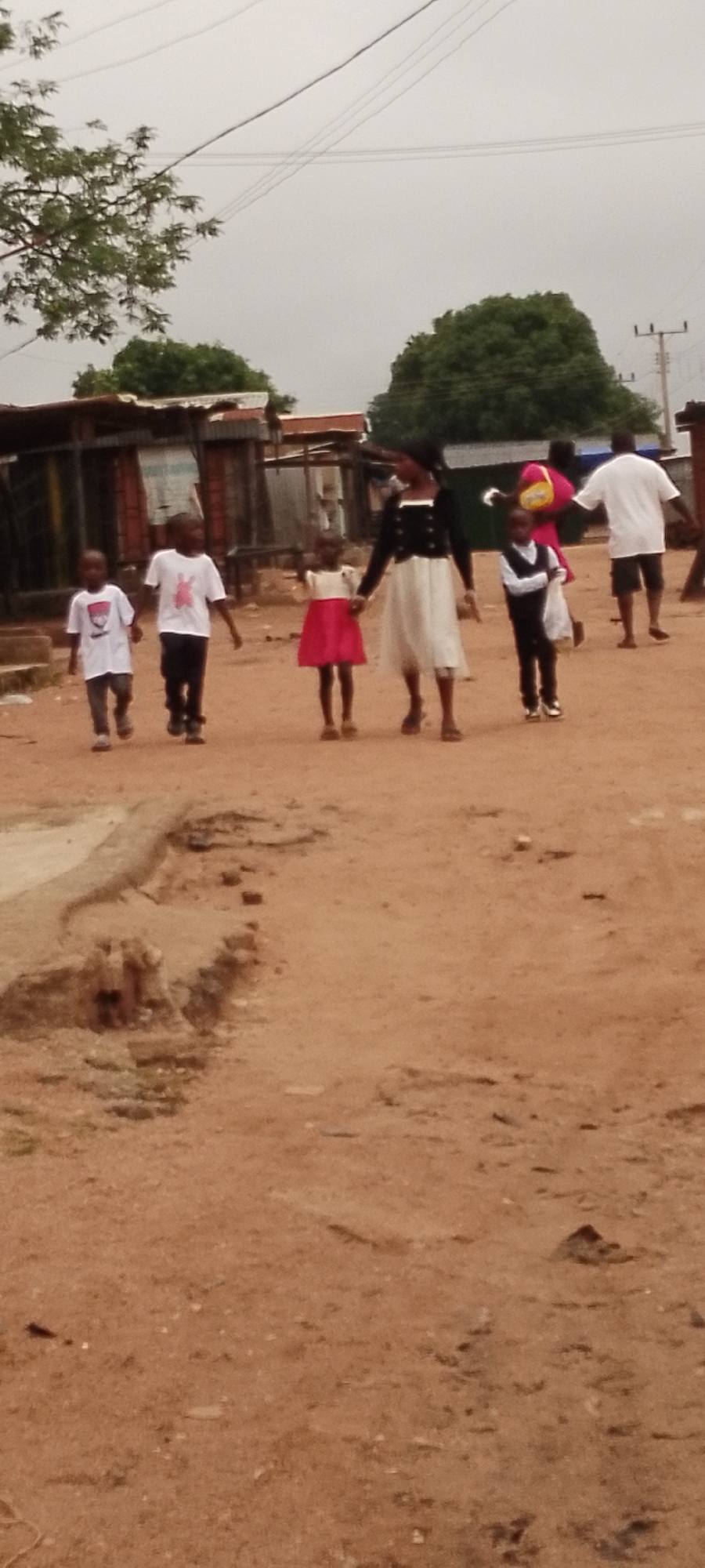 The image depicts a group of people walking on a dirt road in what appears to be a low-income urban or rural area. The sky is overcast. In the foreground, two young boys in white t-shirts and dark pants are walking side by side. To their right, a young girl in a red and white dress and a taller girl or woman in a black and white dress are holding hands. Further in the background, another child in a black vest and white shirt, and two adults, one in a pink garment and another in a white t-shirt and shorts, are visible. The environment is characterized by rough, sandy terrain, with makeshift structures and corrugated metal roofs visible in the background, suggesting a developing or informal settlement. Electrical wires are strung across the sky.