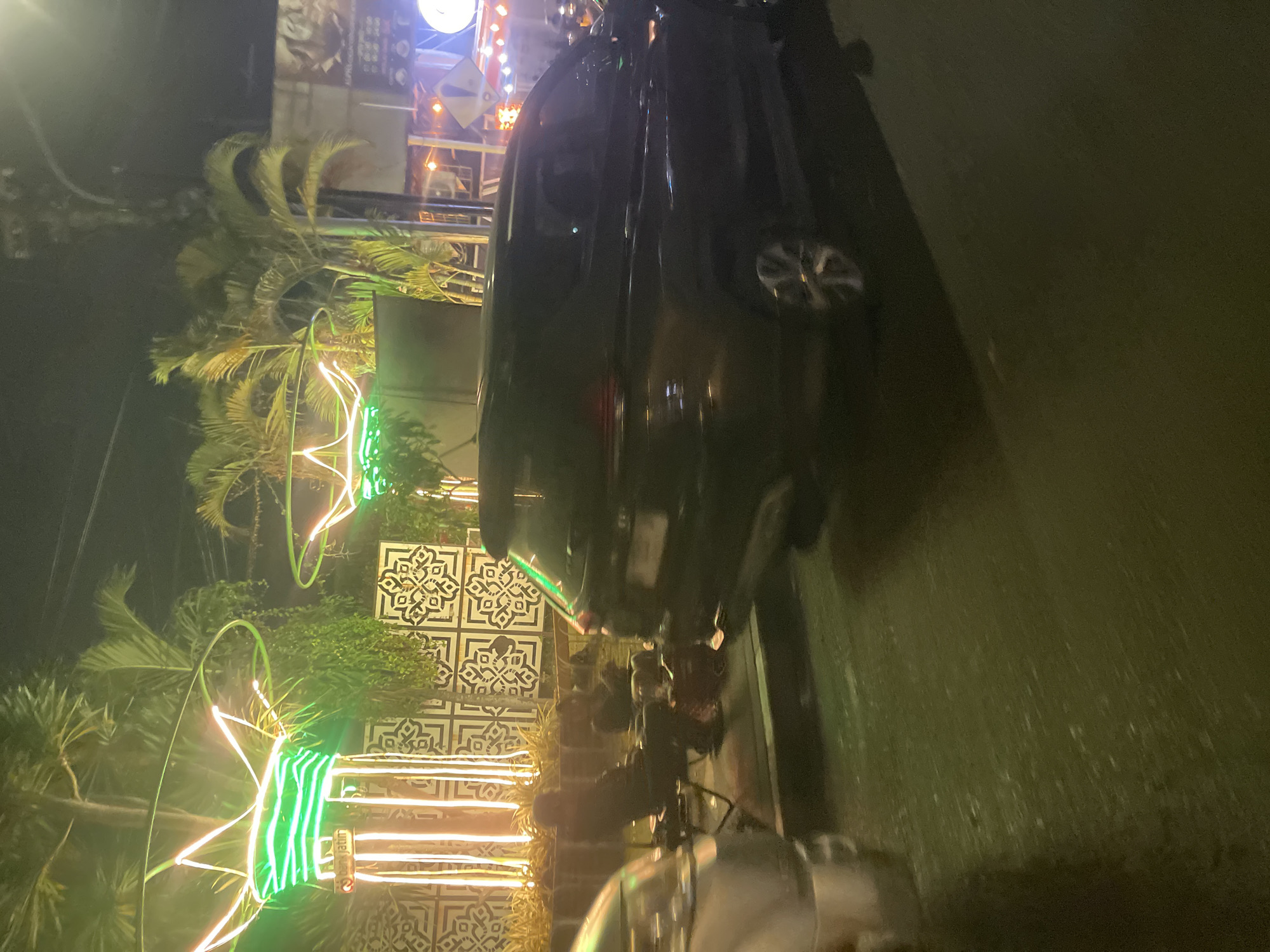 The image captures a nighttime scene likely on a street or sidewalk in Sidoarjo, Indonesia, illuminated by festive green neon lights. In the foreground, a dark-colored SUV is parked, partially obscuring the view of two individuals seated in foldable chairs. The individuals, dressed in dark clothing, appear to be casually observing their surroundings or interacting with each other. Behind them, a patterned wall serves as a backdrop, adorned with vertical light fixtures. Palm trees are visible in the background, adding to the tropical ambiance. The ground is wet, suggesting recent rain or a damp environment. The overall impression is one of a lively evening setting, possibly a commercial or recreational area in Sidoarjo.
