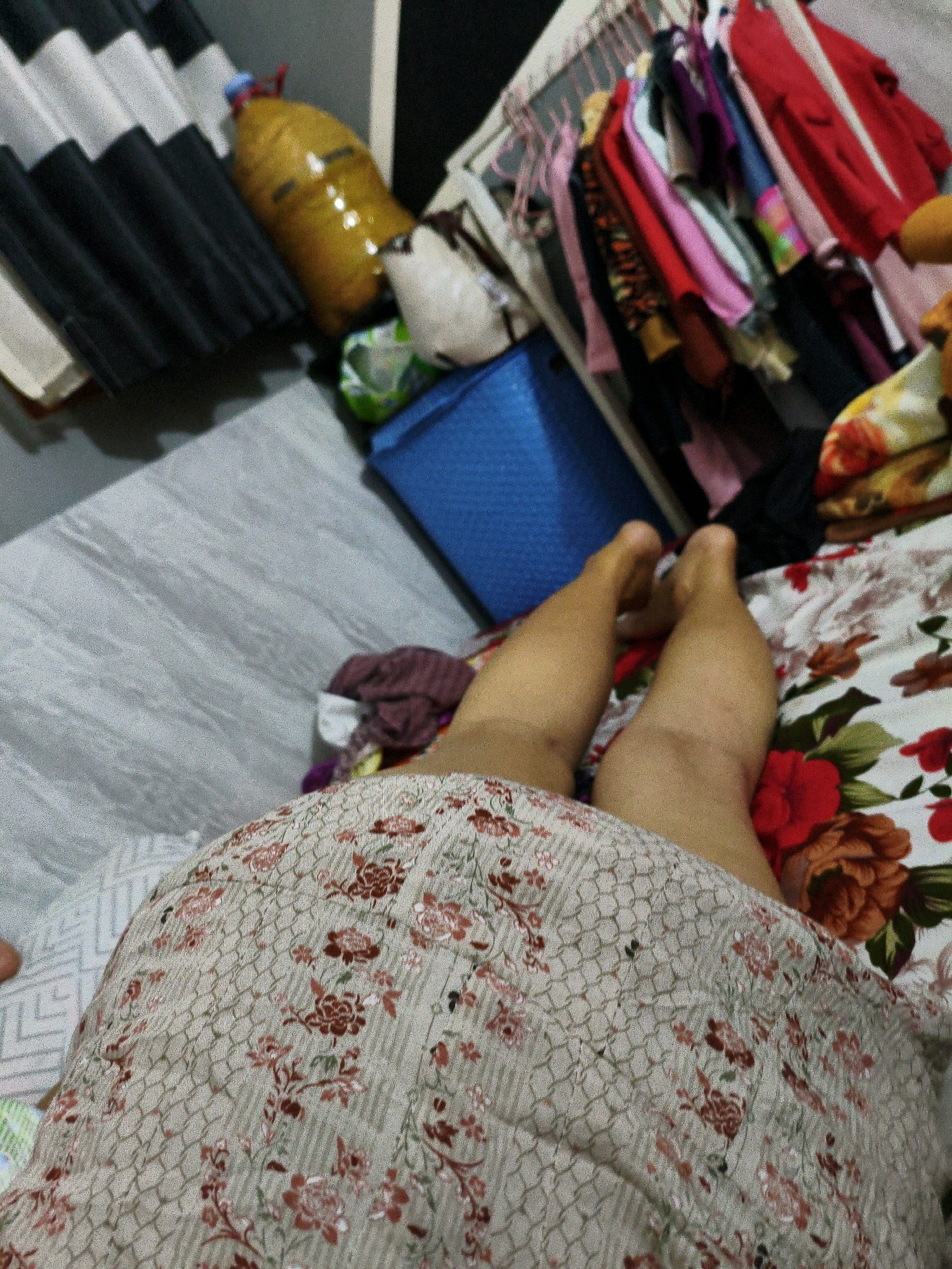 The image depicts a cluttered domestic interior, likely a bedroom or living area, with a person's legs visible in the foreground. The legs are resting on a floral patterned bedspread. In the background, a rack of clothing is visible, along with a large yellow bottle and a blue storage container. The floor is a light gray with a wood-grain pattern. The scene suggests a casual or informal setting. The exact location in Karangtalun, Indonesia cannot be determined from the visual information provided.