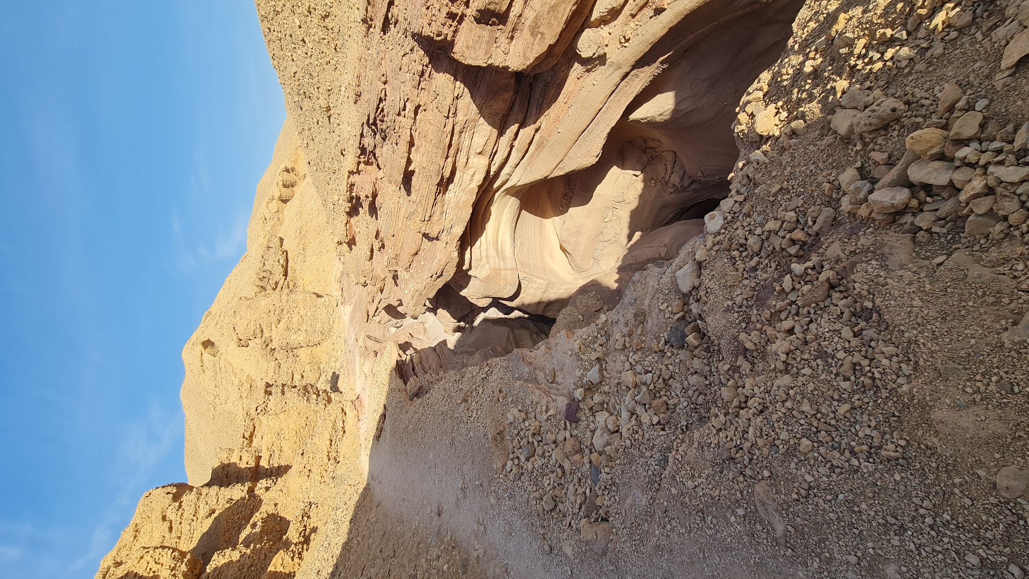 The image shows a narrow, winding slot canyon carved into light brown and beige rock formations. The canyon walls are smooth and sculpted, likely by water erosion. The bottom of the canyon is not fully visible, appearing dark and deep. Loose rocks and gravel cover the edges of the canyon, suggesting an unstable and arid environment. The sky is visible in the upper left corner, a clear bright blue. This is located in the city of Hevel Eilot Regional Council, Israel.