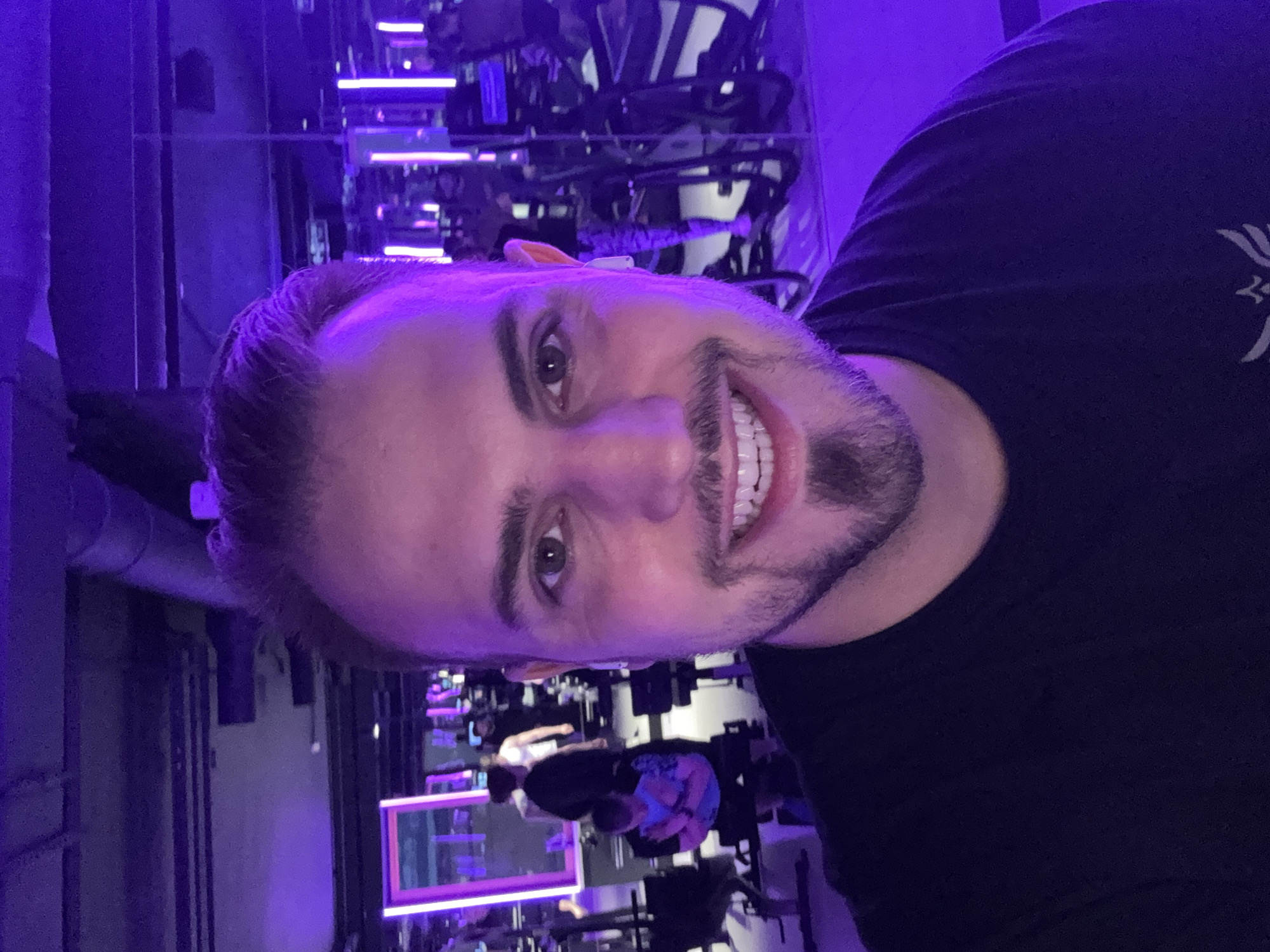 The image is a selfie of a man with short brown hair and a beard. He is wearing a black t-shirt and smiling. The background is a gym with rows of stationary bikes and purple neon lights. Other people are visible in the background, cycling on the bikes. The location is Moscow, Russia.