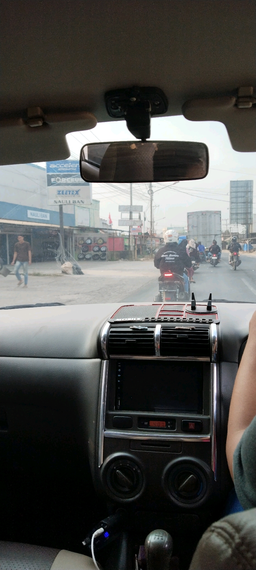 The image is captured from inside a vehicle, looking forward through the windshield and rearview mirror. The scene depicts a street in Yukum Jaya, Indonesia, characterized by a mixed environment of commercial establishments and traffic.
In the foreground, the interior of the car is visible, including the dashboard with an infotainment system, air vents, and a gear shift lever. A person's arm and shoulder are visible on the right side of the frame, suggesting the photographer is a passenger.
Outside the vehicle, the street is bustling with activity. Several motorcycles are in motion, with riders wearing helmets. A man is walking on the sidewalk on the left. Commercial buildings line the street, with visible signage including "ACCELER," "FORCEUM," "ZEETEX," and "NAULI BAN," indicating businesses related to automotive parts or services. Tires are displayed outside one of the shops. In the background, larger vehicles, including a truck, are also present on the road. The overall atmosphere appears to be a typical day in a developing urban or suburban area in Indonesia.
