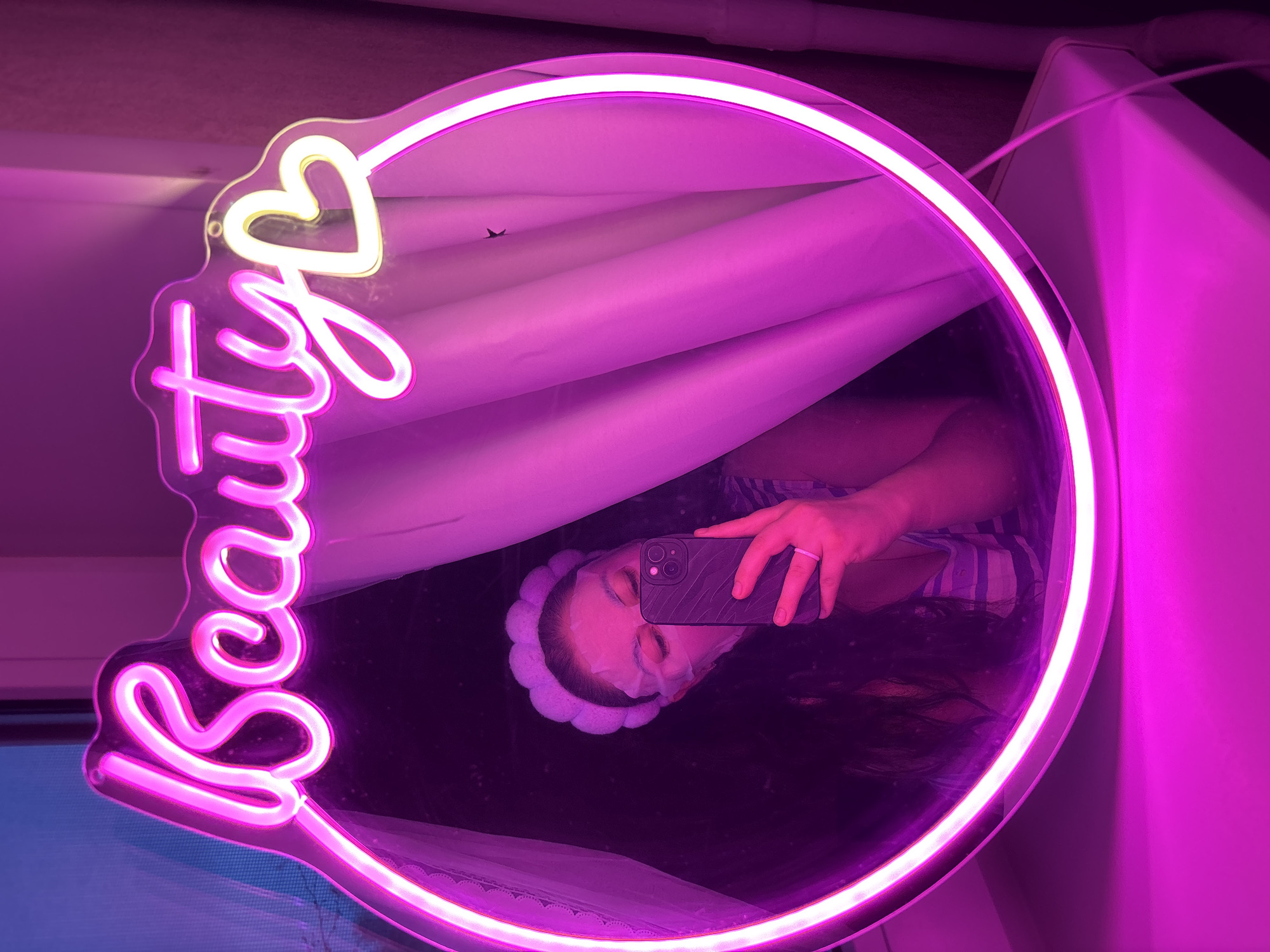 The image shows a mirror with a neon sign reading "Beauty" attached to its upper-left corner. The mirror reflects a person holding a smartphone, possibly taking a selfie. The person is wearing a headband and appears to have a facial mask on. The environment appears to be indoors, potentially a bathroom. The location is stated to be in the city of Chaikovsky, Russia, but there is no contextual information visible in the image to confirm this. The lighting in the scene is dominated by pink and purple hues, likely due to the neon sign and other light sources.