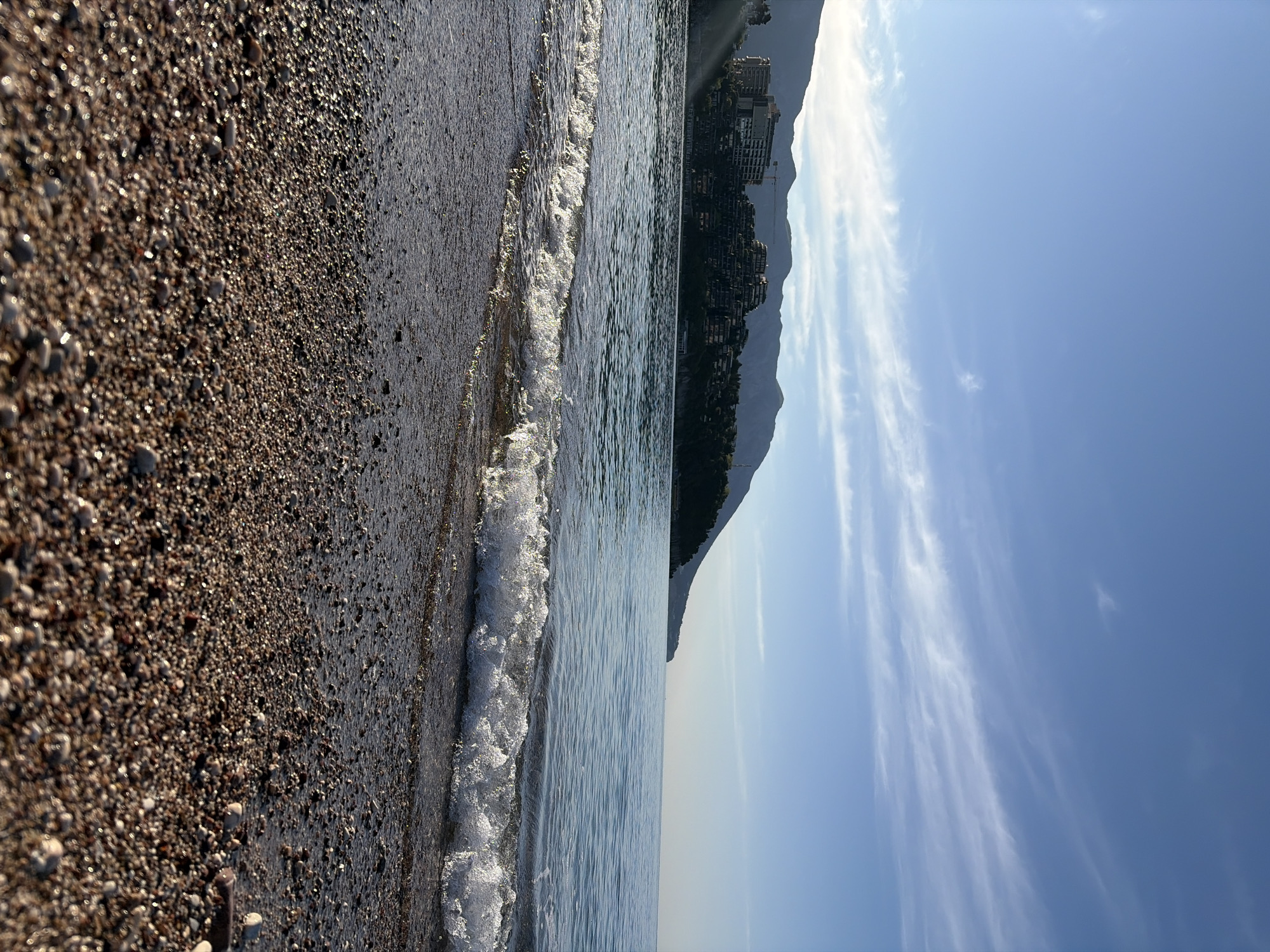 The image shows a beach in Budva, Montenegro, taken from a ground-level perspective with a vertically-oriented composition. The foreground features a pebble beach transitioning into a body of water with small waves. On the right side of the image, across the water, a hill with a visible structure is present under a partly cloudy blue sky.