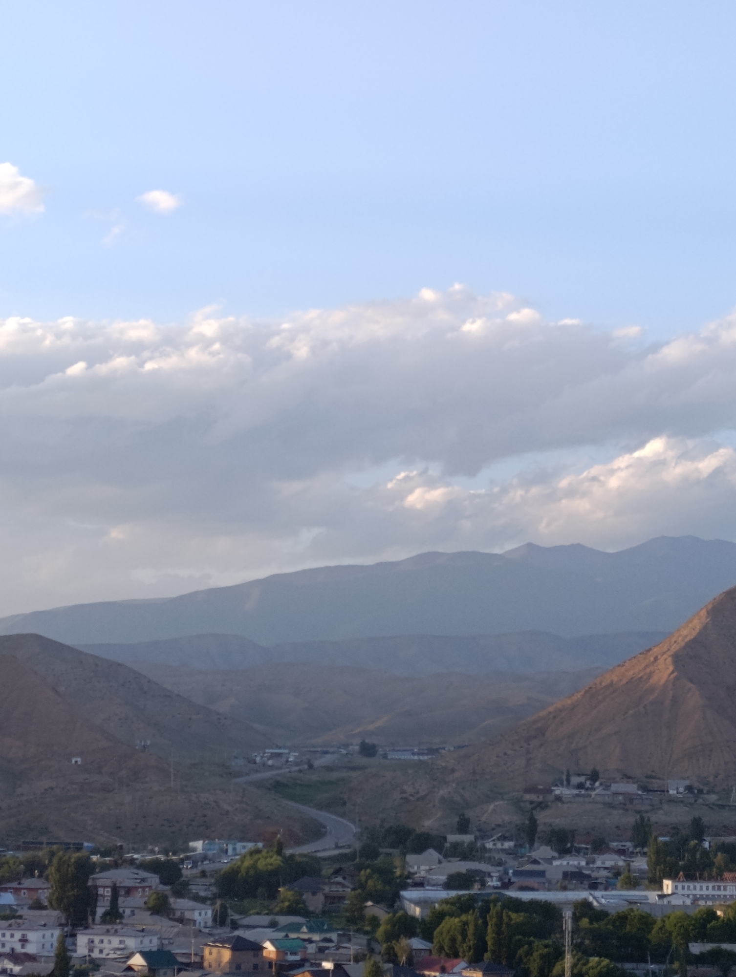 This media file depicts a sprawling landscape at dusk, with a small town nestled in a valley below rolling, arid mountains. The sky is a soft blue, adorned with streaks of white and gray clouds, suggesting fair weather with some cloud cover. The overall mood is peaceful and serene, amplified by the soft lighting of the approaching evening.
The foreground is dominated by the town, a collection of low-rise buildings with predominantly gray and white facades, interspersed with lush green trees. Some buildings appear to be residential, while others might be commercial or public structures, including one with a long row of windows that could be a hotel or apartment complex. A paved road winds through the town and into the valley, hinting at connectivity and a sense of community.
Behind the town, the landscape transitions to the characteristic barren, brown hills of a mountainous region. The terrain is characterized by gentle slopes and gradual inclines, with layers of mountains receding into the distance. The light casts long shadows, highlighting the rugged contours of the land. One prominent mountain peak in the right of the frame is illuminated by the warm, golden light of the setting sun, creating a striking contrast with the shadowed valleys.
The time of day is clearly dusk, with the sky exhibiting the soft, diffused light typical of this period. The location cues, such as the arid mountain terrain and the style of the buildings, are consistent with a settlement in Central Asia, specifically Naryn City, Kyrgyzstan, as suggested by the provided context. No people are visible in the media file, and no text is discernible. The scene is a quiet, natural vista, capturing the tranquil beauty of a town at the foot of imposing mountains as day turns to night.