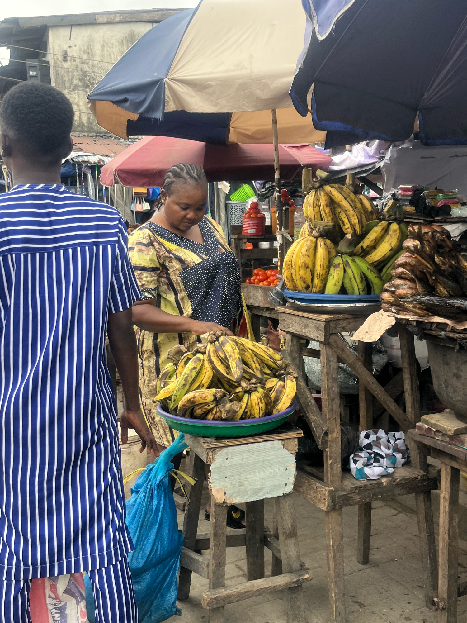 A woman, dressed in a yellow and black patterned dress, stands behind a market stall in Lekki Phase I, Nigeria. She is attending to a large bowl of green and yellow bananas. To her left, a person wearing a blue and white striped shirt stands with their back to the camera. The market stall is made of wood and features a variety of goods including more bananas, red tomatoes in a white basket, and stacked fish. Two large umbrellas, one blue and white, and another brown and red, provide shade over the stall. The ground is paved and the background shows buildings and other market stalls.