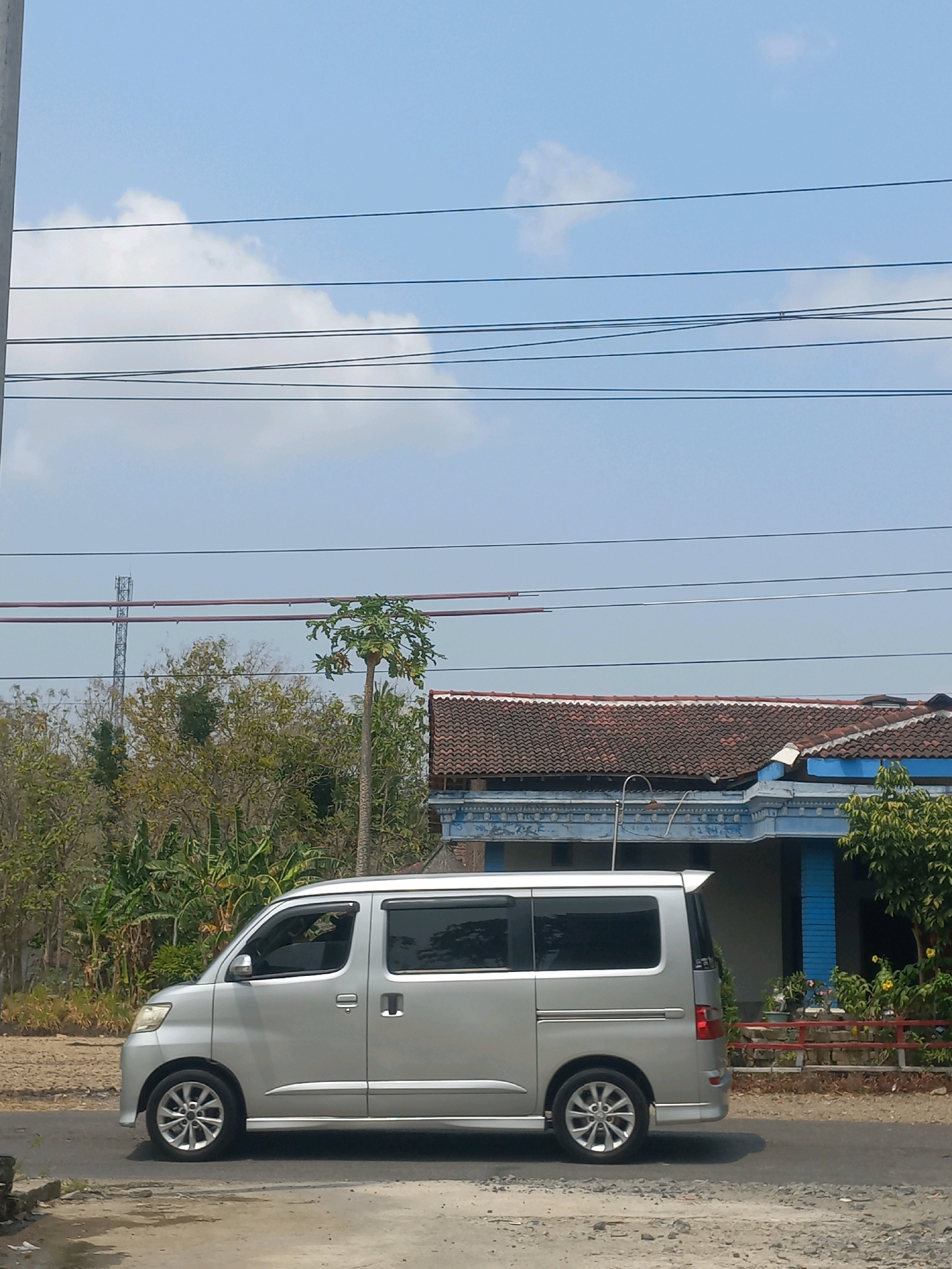 A silver minivan is driving on a road in Baturetno, Indonesia. The minivan is seen from the side, and it is silver in color. The road is paved, and there are some trees and a house in the background. The sky is blue, and there are some clouds in the sky. The minivan has tinted windows and a spoiler on the back. The house in the background has a red tile roof and blue walls. There is also a papaya tree in the background. The image was taken during the day, and the lighting is bright.
