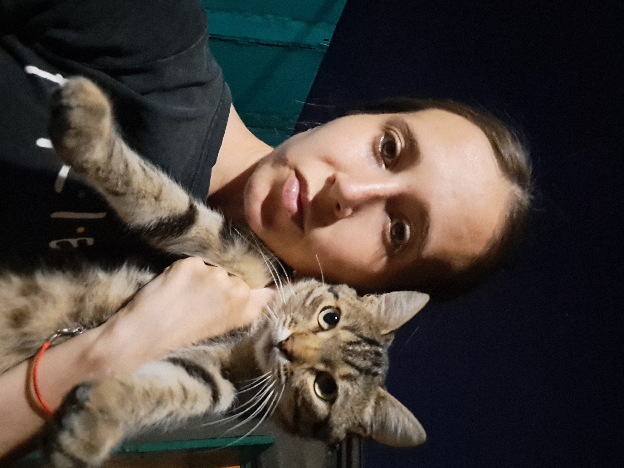 A young woman with light brown hair, wearing a black t-shirt, holds a tabby cat in front of her chest. She is looking at the camera with a neutral expression, and her mouth is slightly open. The cat is looking towards the camera with yellow eyes. They are standing outdoors at night, with a dark blue sky in the background and a teal fence to the left. The woman has a red bracelet on her left wrist. The image is a selfie, taken from a slightly low angle.