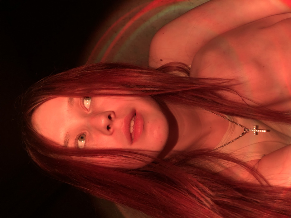 A close-up portrait features a young female subject illuminated primarily by a strong red-orange light. Her long, reddish-brown hair frames her face and extends over her shoulders. Her light-colored eyes are directed towards the upper right of the frame, and her lips are slightly parted. She is wearing a light-colored garment, likely a top, and a silver-toned chain with a small, ornate cross pendant is visible around her neck. Her right shoulder and a portion of her upper arm are visible in the lower right, positioned as if embracing or resting against another object or person. The background is largely dark and out of focus, with faint, arcing red light patterns visible on the right side. The visual evidence within the image does not provide specific details to identify the location as the city of Фоки, Russia; the environment appears to be an indoor setting with controlled, colored lighting.