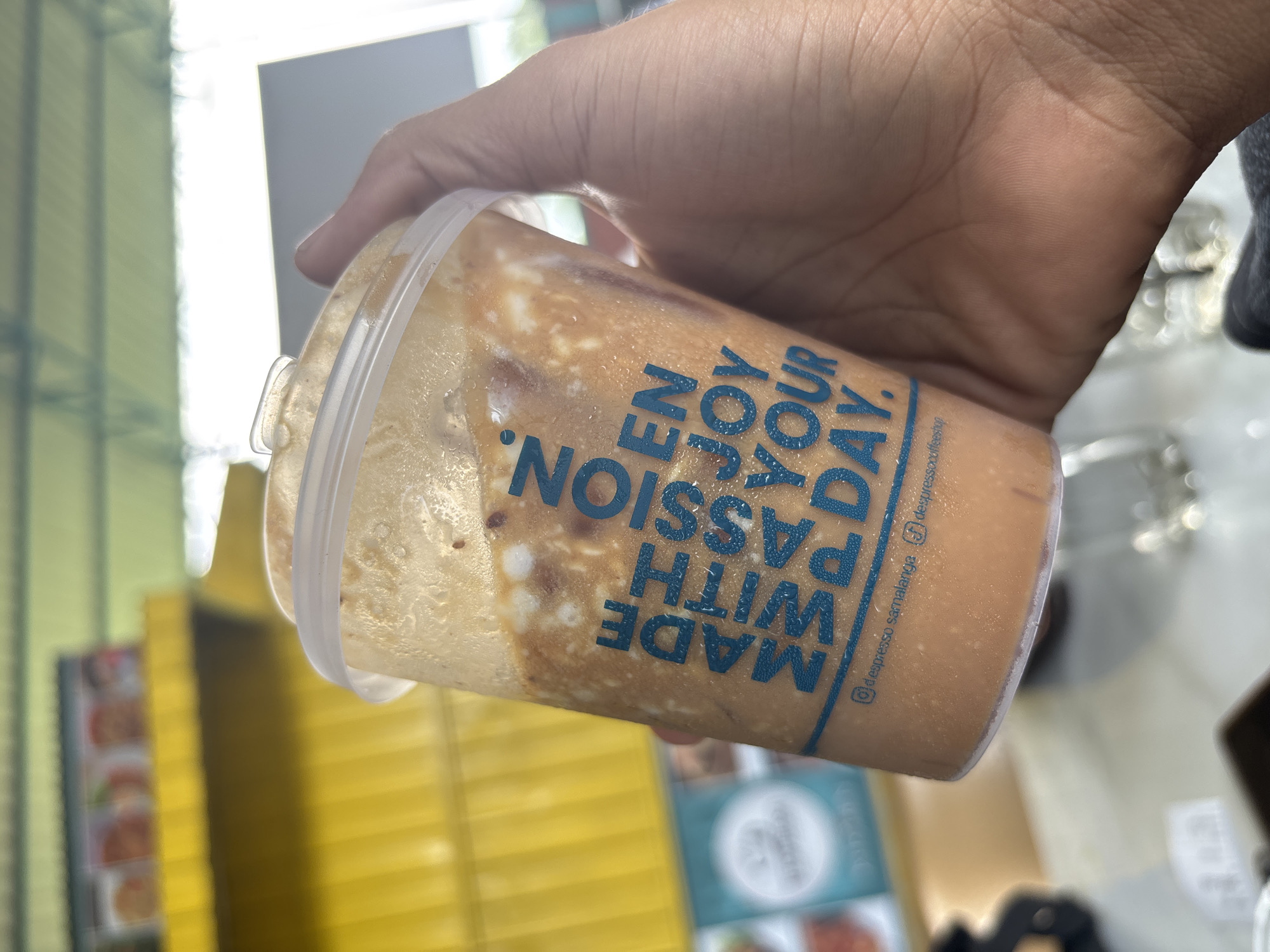 A person's hand holds a clear plastic cup containing a cold beverage, likely an iced coffee or tea, characterized by light brown liquid and swirling white, creamy components. Visible condensation on the outside of the cup indicates its chill. Blue text is printed vertically on the cup, reading "ENJOY YOUR DAY." and "MADE WITH PASSION." Below these phrases, smaller text appears to be social media handles: "@despresso_samalanga" and "@despressocoffee".
The scene is set outdoors or in a semi-outdoor commercial environment in Namplohbaro, Indonesia, under bright daylight. The background is blurred, showing yellow vertical slats, possibly from an awning or a building facade, along with indistinct signs and structures. The primary activity depicted is the act of holding the refreshing drink. The bright illumination suggests it is daytime, and the cold beverage implies warm weather. No specific emotions are directly conveyed by the limited visible subject matter.