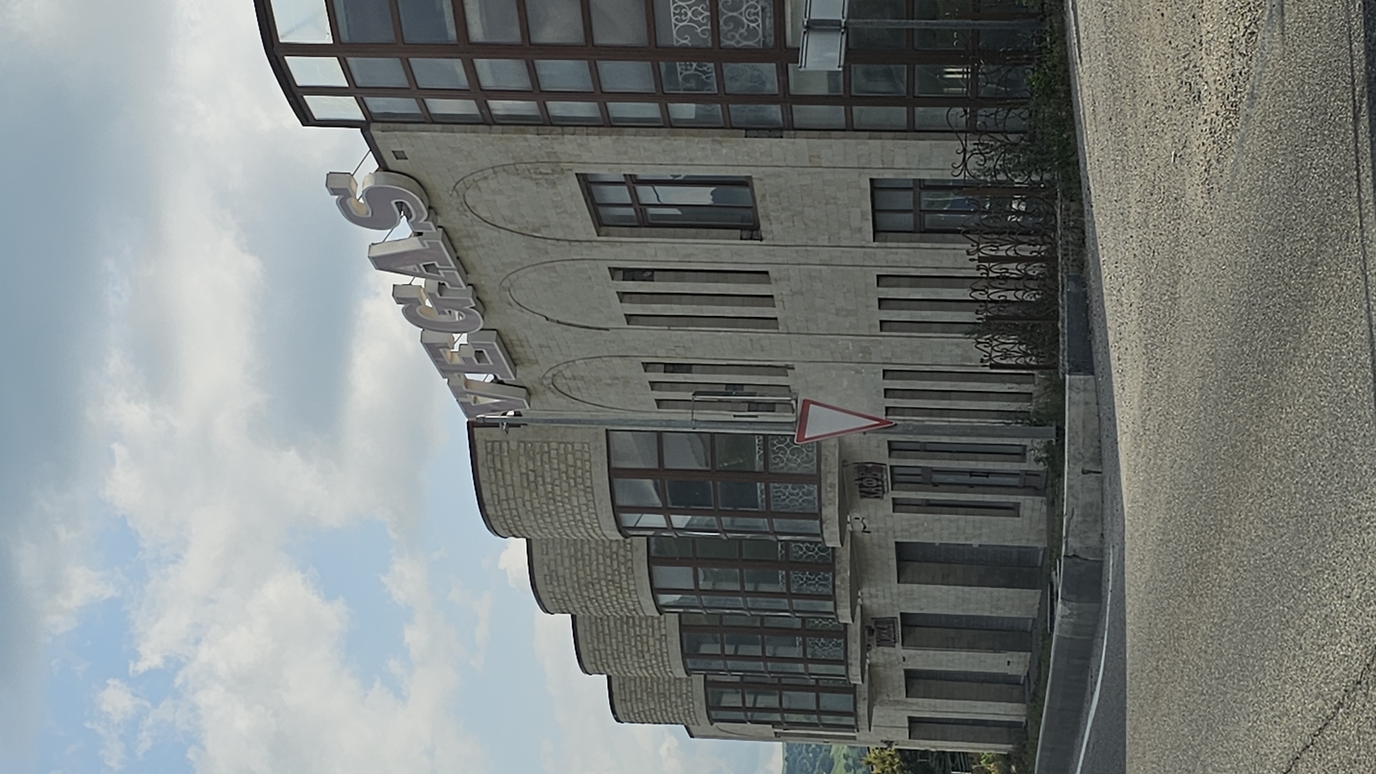 The image depicts the exterior of a building in Podkumok, Russia, with prominent signage spelling out "VEGAS" in large, light-colored letters across the top facade. The building is constructed with light-colored stone or brick and features a mix of architectural styles. On the left, there are several rounded sections with bay windows, each topped with a cylindrical brick structure. The central part of the building has a series of tall, narrow vertical windows set within arched recesses. To the right, a section of the building is clad in darker glass panels with a grid-like frame. A decorative wrought-iron fence runs along the front of the building, partially obscured by greenery. Below the "VEGAS" sign, on the lower level, there appear to be several storefronts or entrances, one of which has signage that seems to read "КАФЕ" (Cafe). A triangular red and white warning sign is visible on the building's facade. The building is situated alongside a paved road, with a curb and sidewalk area in front. The sky above is partly cloudy. No people are visible in the image.