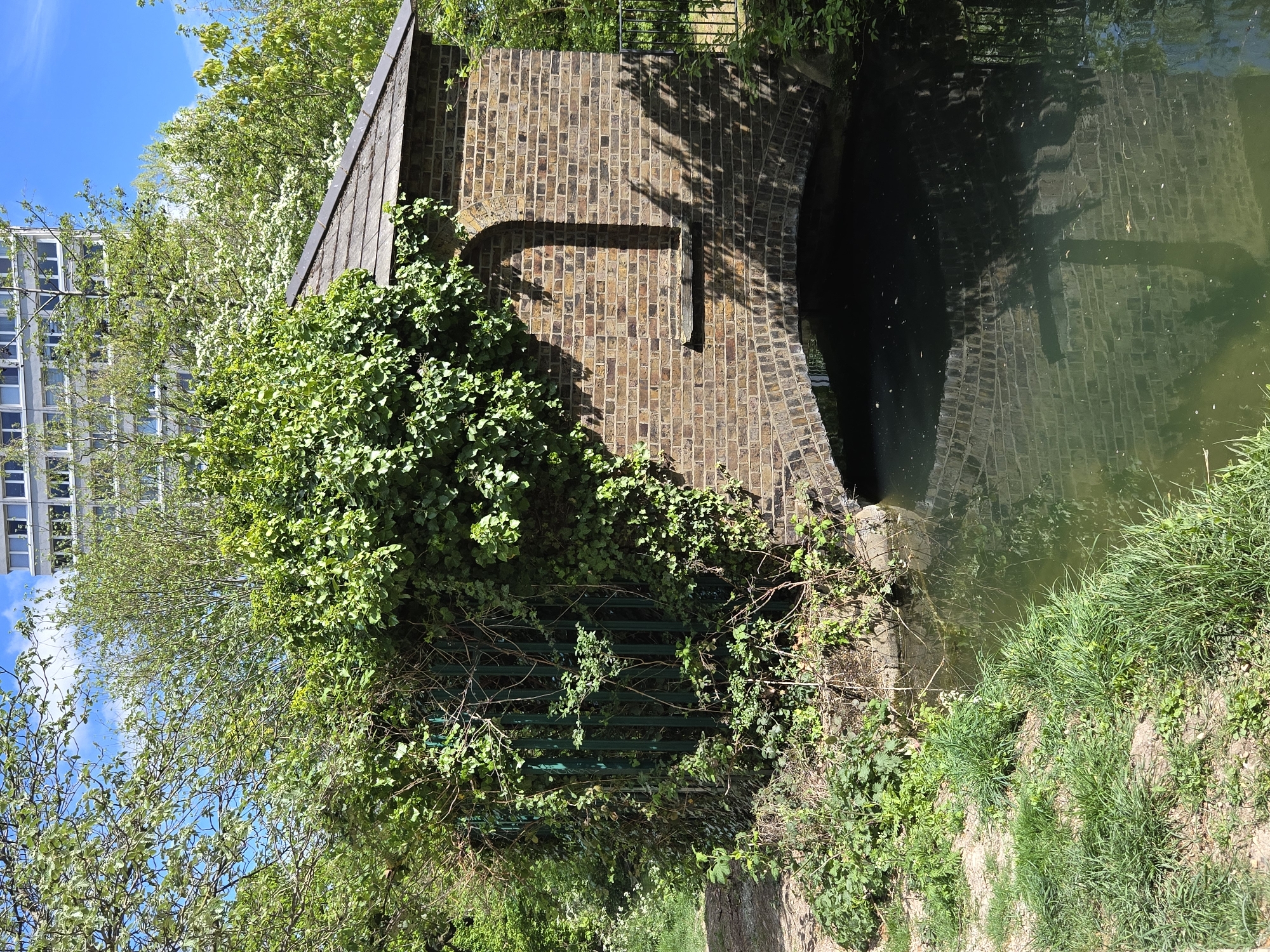This image shows a canal in London, United Kingdom. A small brick building stands on the bank, partially covered in ivy. The building has an arched opening leading to the water. The canal water is murky green and reflects the brickwork and shadows of the building. Lush green grass and vegetation grow along the bank. In the background, a modern apartment building is visible. The sky is blue with scattered clouds.