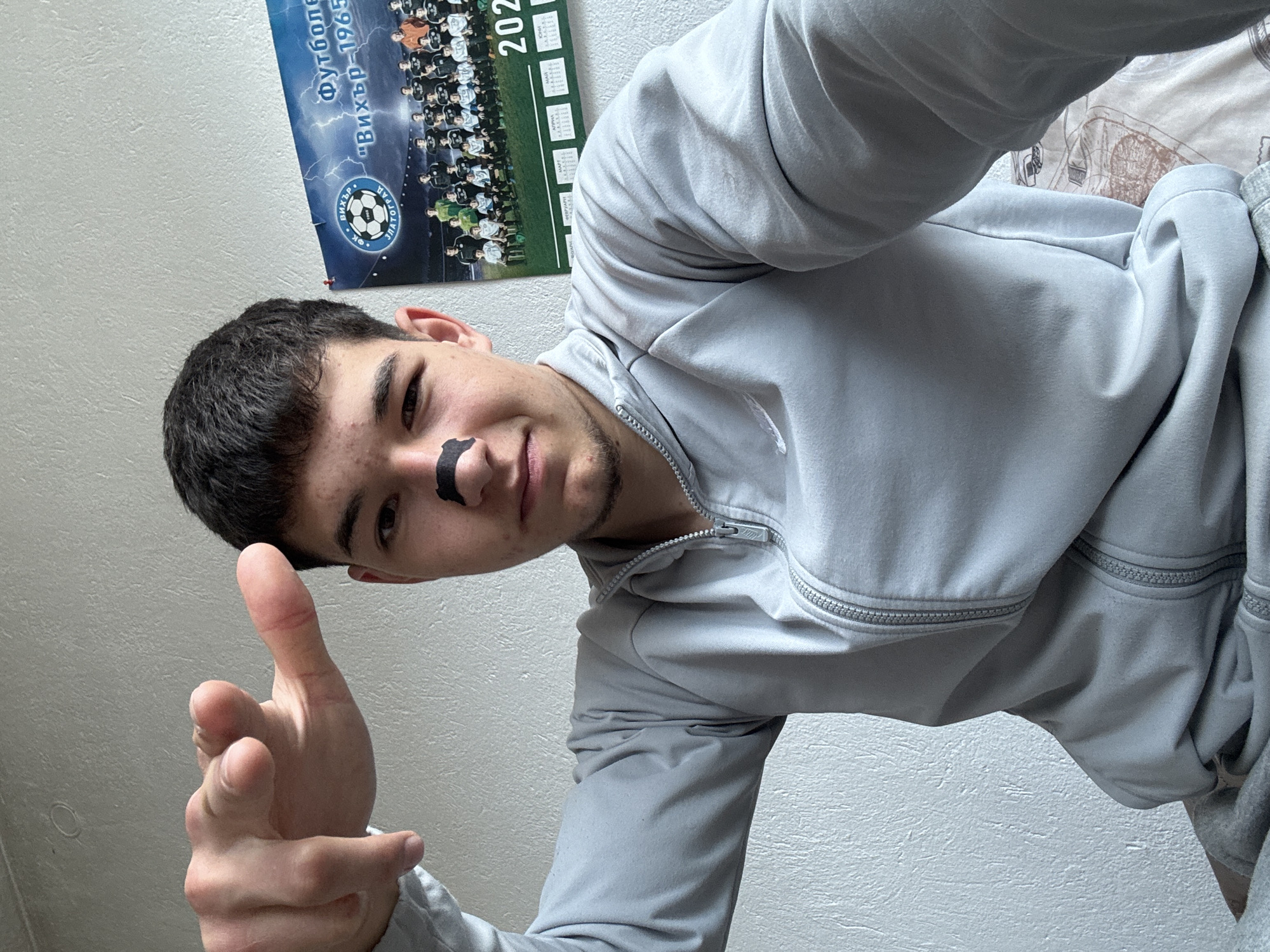 A young man is taking a selfie in a room. The man has short dark hair and a strip of black tape is on his face under one eye. He is wearing a gray zip-up jacket and is holding up his hand, pointing his thumb at the camera. Behind him is a wall with a poster of a soccer team with the words "Футболен клуб 'Вихър-1965'" and "2023" on it. The text indicates the location is likely Zlatograd, Bulgaria, where the soccer team FC Vihur-1965 is based.