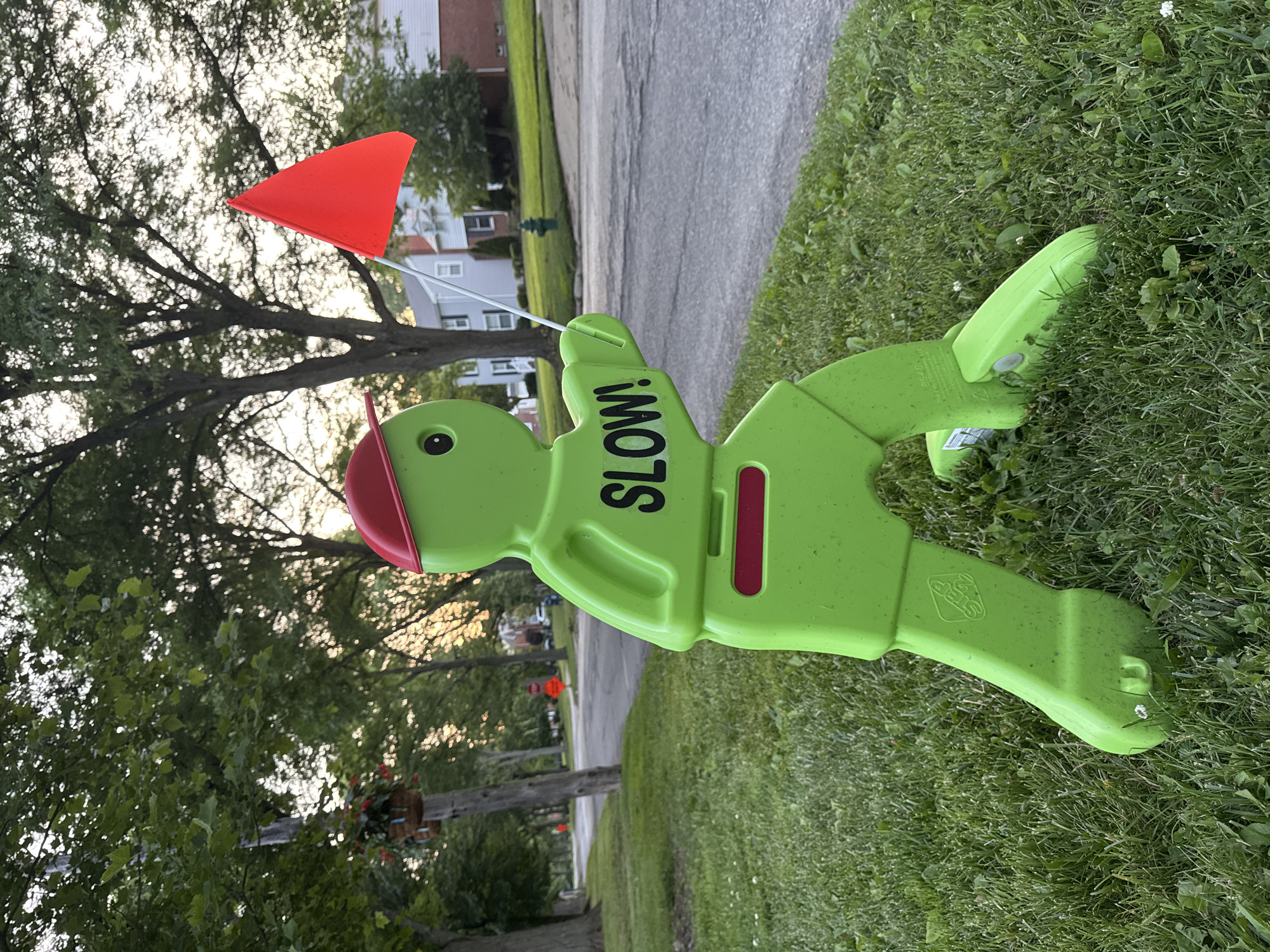 An eye-level, medium shot captures a bright green, plastic lawn figure with a red hat and a red flag in its right hand. The figure is positioned on the left side of the frame, standing on a lawn with green grass and scattered white flowers. Black lettering on the figure's torso reads "SLOW!". The figure is a simplified human-like shape with oversized limbs and a round head with one visible eye. Behind the figure, a paved street runs diagonally from the bottom left to the upper right of the frame. Trees with green foliage line the street, and houses with white and grey siding are visible in the background. The sky is overcast with a soft, warm light. The scene appears to be a residential street in Shaker Heights, Ohio, USA.