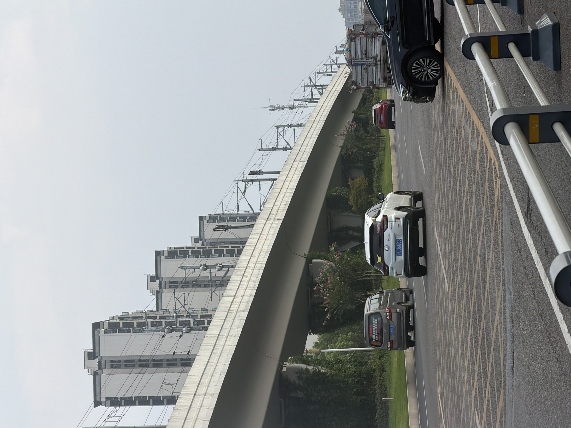 The image shows a section of elevated railway or highway in Jiangjin, China, with several vehicles on the road below. The elevated structure is a long, concrete bridge supported by pillars. Overhead, there are numerous electrical wires and poles associated with the railway or monorail system. In the background, several tall, modern apartment buildings with white facades and dark windows are visible, lining the horizon.
On the road, there are at least four vehicles. Closest to the viewer on the right is a black sedan. To its left, a white SUV with a yellow Pikachu sticker on the rear window is driving away. Further left, a gray minivan is also moving away from the viewer. In the middle distance, a red SUV is seen driving away. The road surface is asphalt, and there are visible yellow grid markings and white lane dividers. The environment appears to be a moderately built-up urban area with some greenery, including trees and bushes, visible along the side of the road and supporting the elevated structure. The sky is overcast with a pale gray hue.