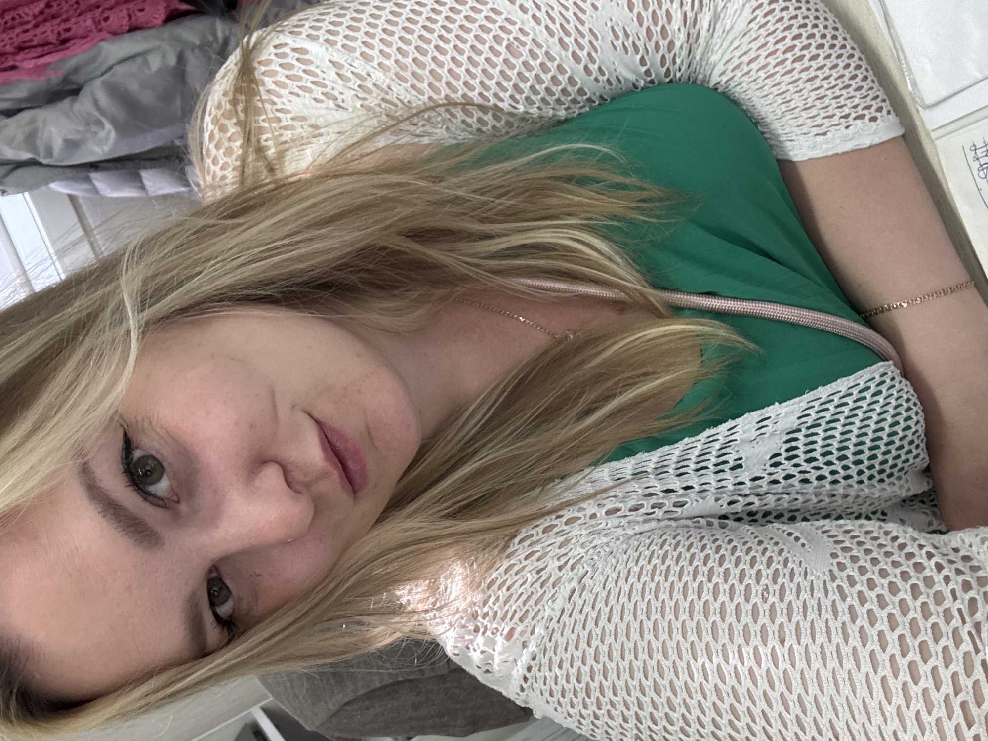 A woman with blonde hair is wearing a green top with a white fishnet-style overshirt. She is wearing a thin gold necklace and a gold bracelet on her right wrist. The background includes a mix of gray and pink fabrics and what appears to be a window with blinds. There is no evidence to suggest the location is in Noviy, Russia.