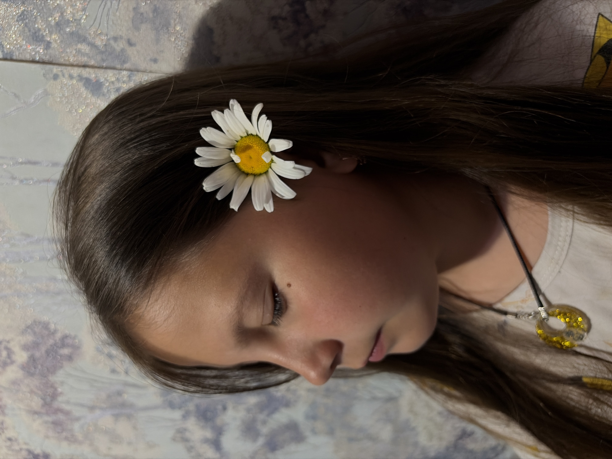 A young person lies on a surface with a floral pattern. They have long, dark hair and a daisy flower is positioned near their ear. They are wearing a white shirt and a necklace with a round pendant. The location is Unknown, Unknown.