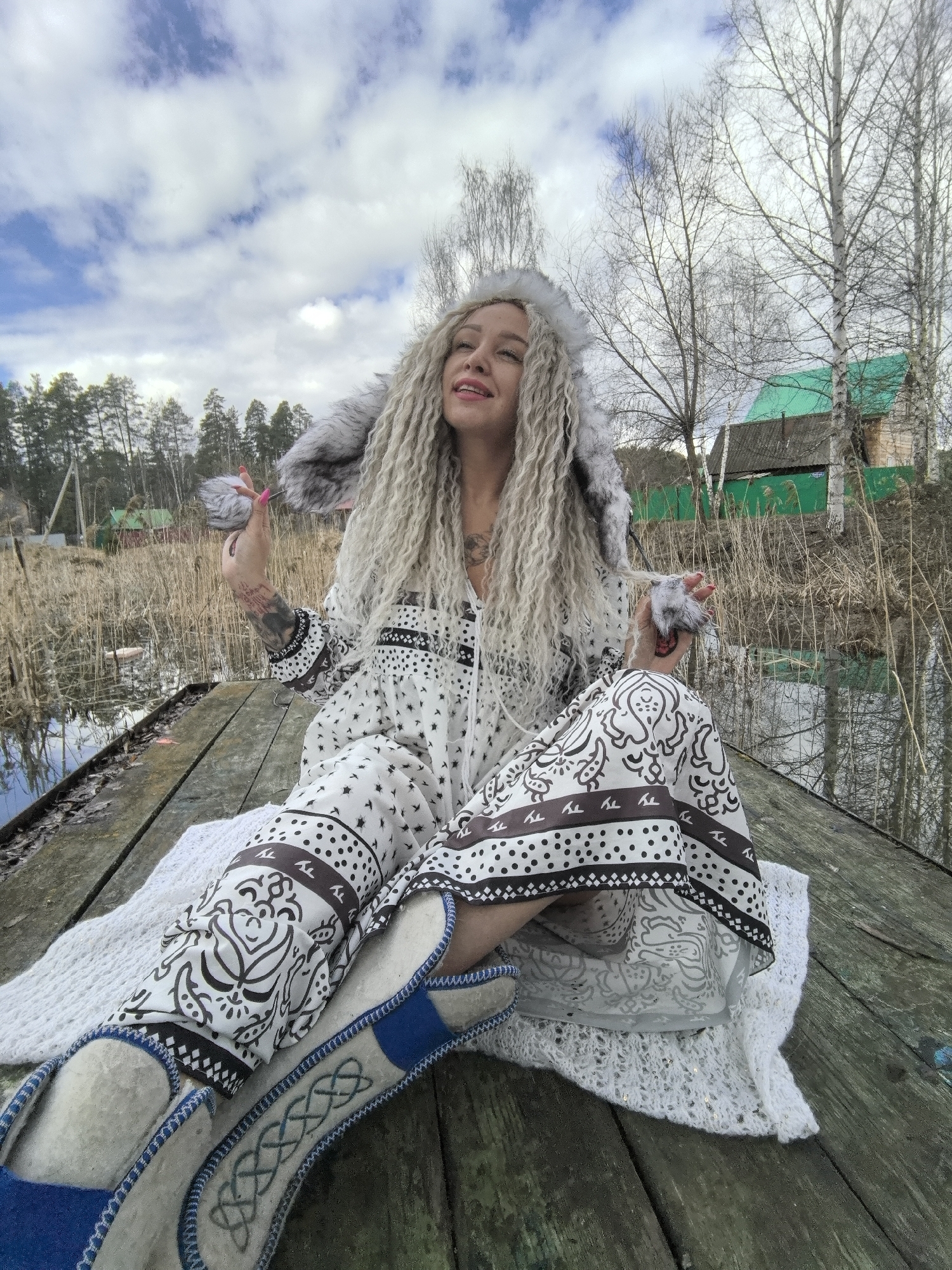 A person is seated cross-legged on a weathered wooden dock. The individual is adorned in a white patterned dress with dark brown trim, a fur-trimmed hooded jacket, and blue-and-white felt boots. The person has long, light blonde dreadlocks. Both hands hold small, furry items. A knitted white blanket lies beneath the person. Reeds grow near the water's edge, and in the background are bare trees and a green-roofed building. The setting appears to be a natural environment with scattered structures, consistent with a location in the city of Noviy, Russia. The sky is cloudy.
