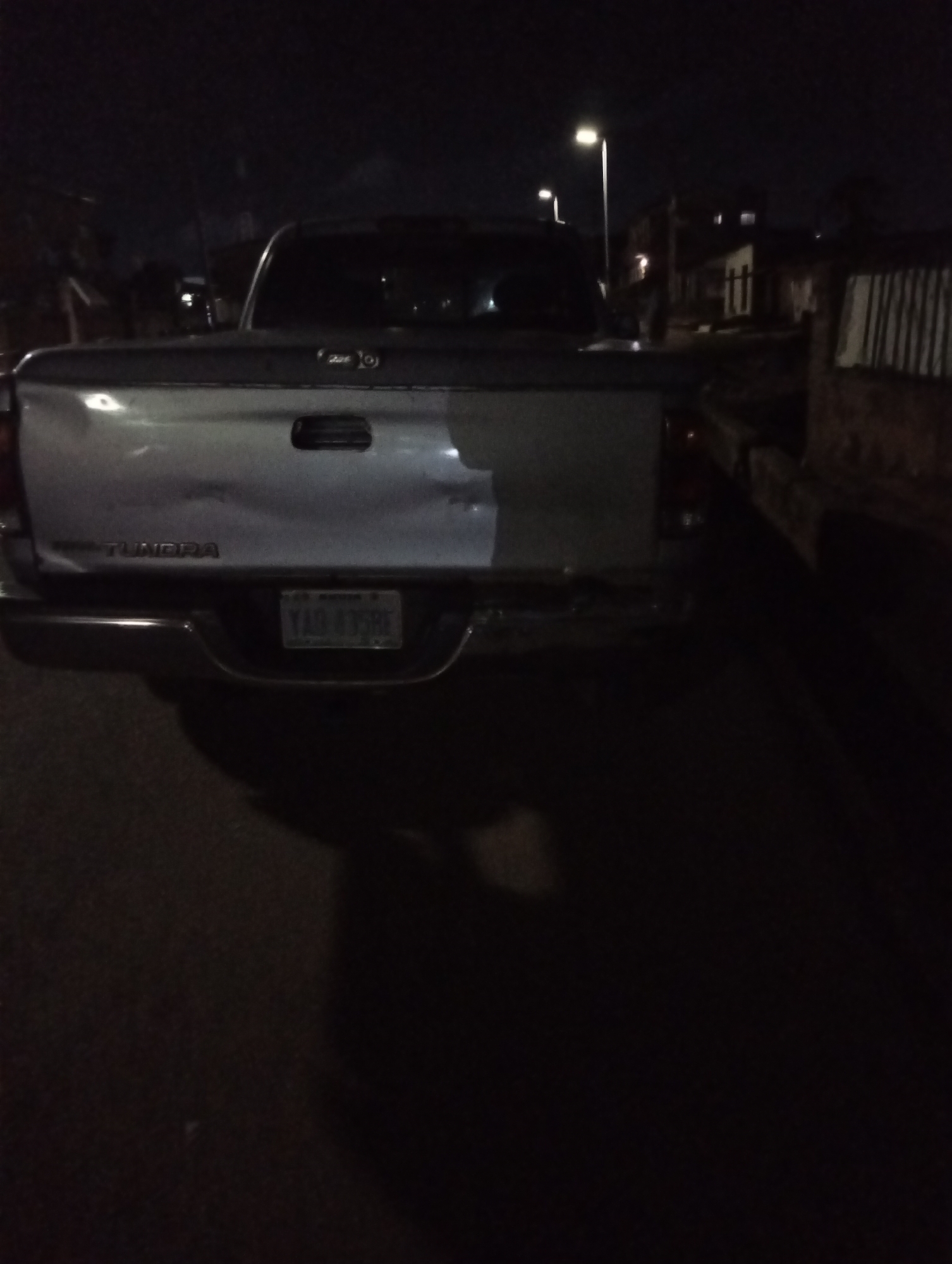 At night, a silver Toyota Tundra pickup truck faces the camera on a dark street. The truck has a dent on the tailgate. A vehicle or trailer is parked to the right of the Tundra. The license plate is visible and reads "YAR 895RE." Streetlights illuminate the scene. It is not possible to confirm the location of Jos, Nigeria with the provided image.