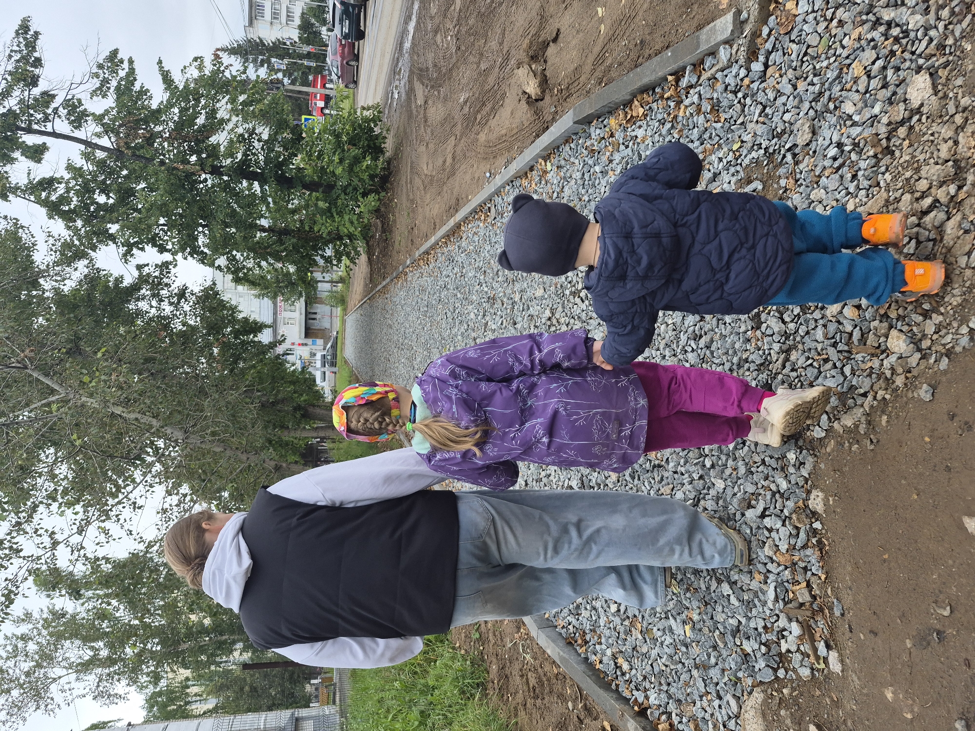 The image shows three people walking away from the camera on a gravel path. The person in the lead is an adult wearing a white hooded sweatshirt under a black puffer vest and light blue wide-leg jeans. To their right, a young girl is walking, wearing a purple jacket with a floral pattern, pink pants, and a colorful knitted hat. She is holding hands with the adult. On the adult's left, a younger child is walking, wearing a dark blue puffer jacket with bear ears on the hood and teal pants, also holding hands with the adult. The path is bordered by a curb and a patch of bare dirt. In the background, there are trees, buildings, a paved road with cars, and a fire truck. The sky is overcast. The scene appears to be in an urban area, possibly a park or a newly constructed walkway. The location is identified as Chaikovsky, Russia.