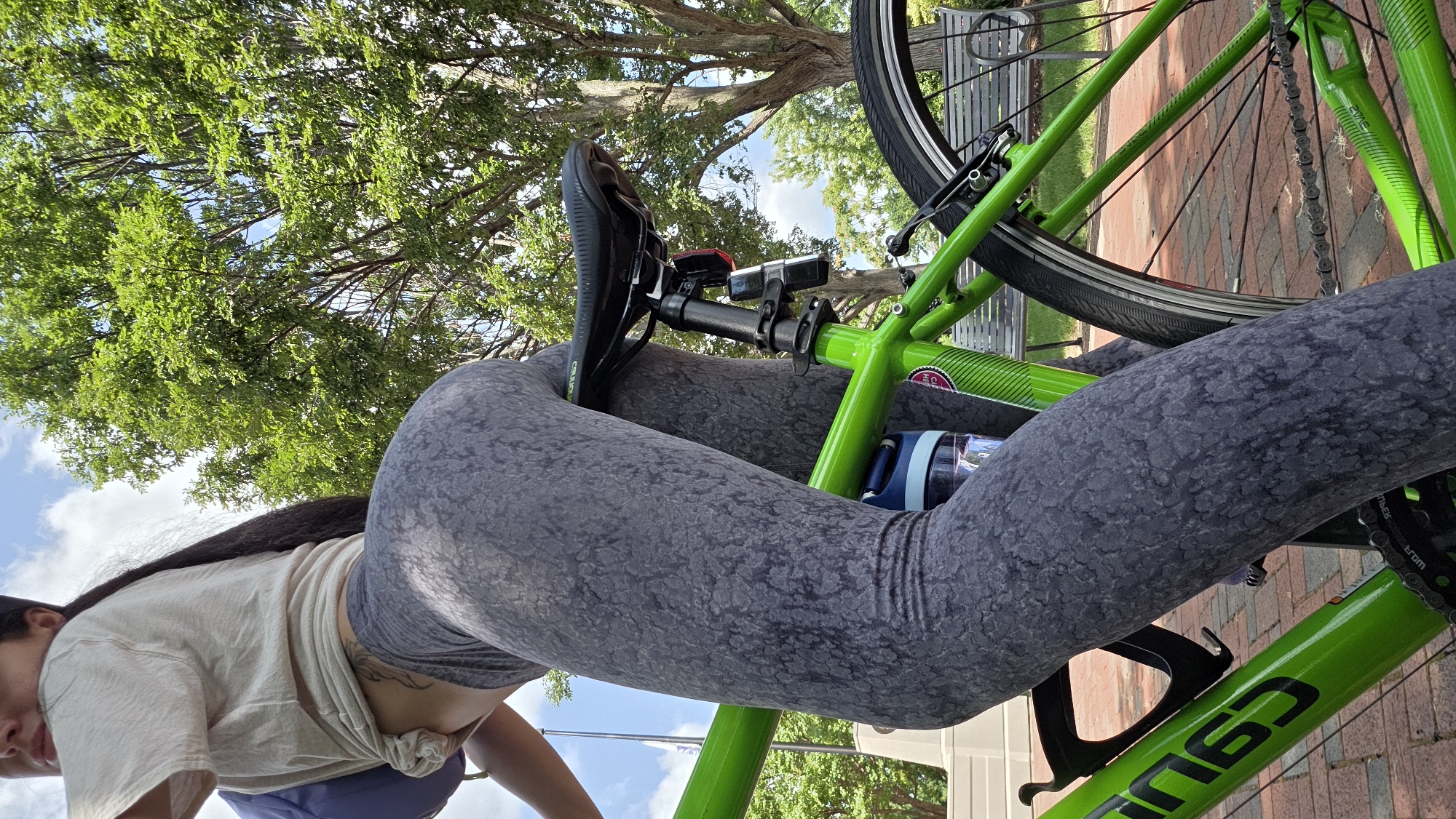 A person is positioned on a bright green bicycle, with their lower body and torso prominently featured. They are wearing a light-colored t-shirt tied at the waist and dark grey patterned leggings. The bicycle has a black seat, a water bottle attached to the frame, and visible chain and pedals. The scene appears to be outdoors, with lush green trees and a blue sky with white clouds in the background. The ground is paved with red bricks. There is a white monument or pillar visible in the background to the left, and a park bench to the right.
