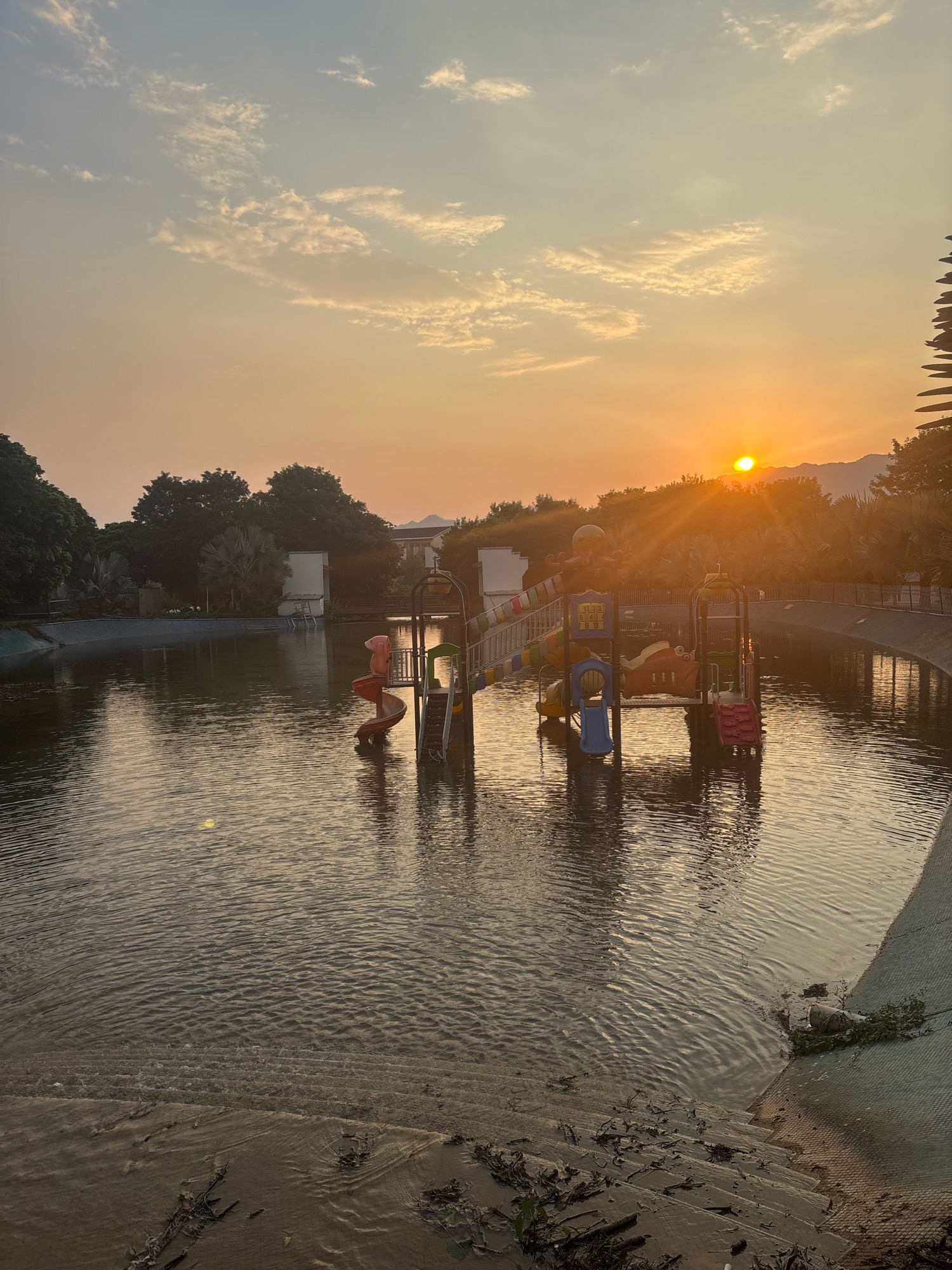 The image depicts a sunset over a body of water, likely a man-made pool or shallow lake, situated in the city of Sanshui District, China. The water's surface is rippled, reflecting the warm hues of the setting sun. In the middle of the water, a small water park structure is visible, featuring colorful slides and climbing elements, including a red spiral slide and a yellow and blue multi-level play area with animal-themed decorations. Trees line the perimeter of the water, and in the distance, mountains are silhouetted against the orange and yellow sky. The sun is low on the horizon, casting long rays of light that fan out across the sky and water. The foreground shows a sandy, stepped embankment leading into the water, with some debris scattered along the edge. The scene appears serene and uninhabited, with no people visible.