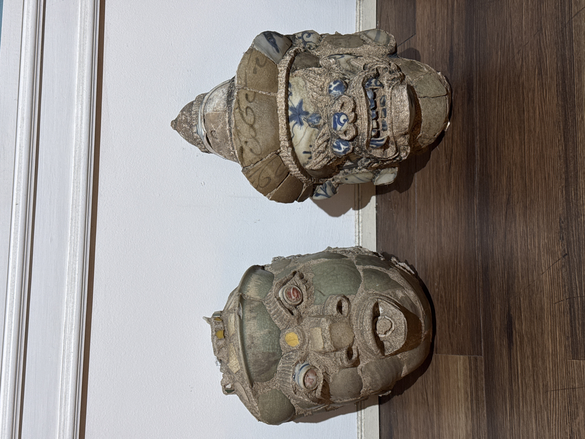 Two weathered, mosaic-style masks, one depicting a human-like face and the other a more monstrous visage, stand on a dark wooden floor against a white wall with decorative molding. The mask on the left has green and yellow ceramic pieces forming its face, with red and blue glass for eyes. The mask on the right is adorned with blue and white patterned ceramic shards, resembling a warrior's helmet. Both masks appear to be made of a rough, porous material, possibly clay or concrete, with visible cracks and missing pieces. The lighting casts shadows on the wall behind the objects. The image is taken from a medium distance, showing the full height of the masks and a portion of the floor and wall. The location is Samrong Nuea Subdistrict Municipality, Thailand.