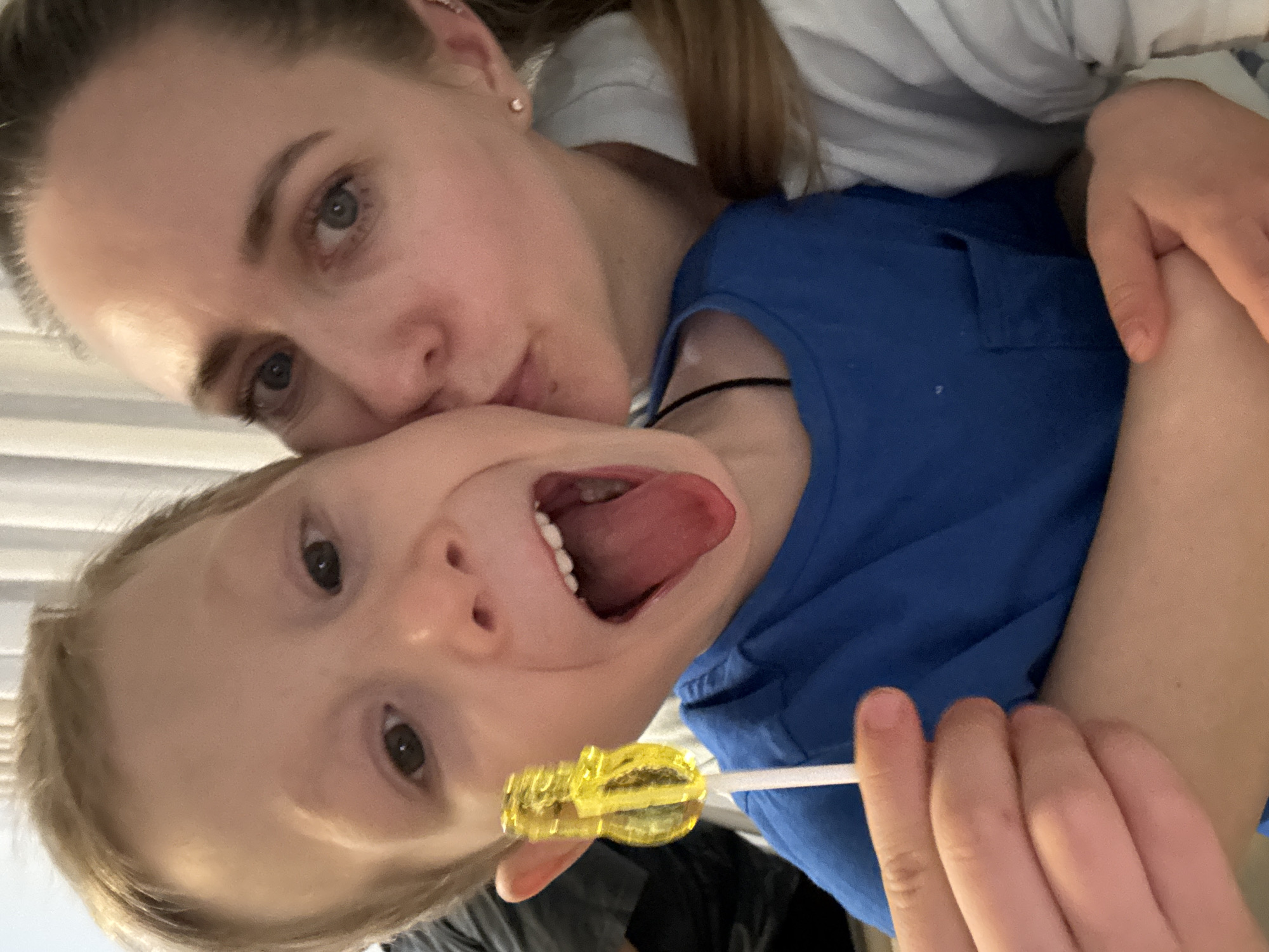 A fair-skinned adult female with light brown hair is positioned behind and partially to the side of a fair-skinned child with blond hair. The woman is giving the child a kiss on the cheek. The child is sticking their tongue out and holding a yellow lollipop. The child is wearing a blue shirt and the woman is wearing a white shirt. The location is unknown.
