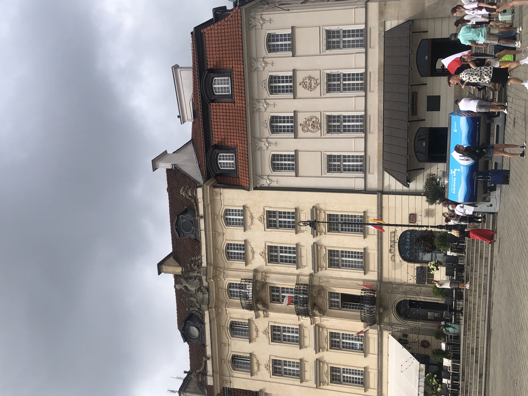 The image shows a street scene in Warsaw, Poland, likely in the Old Town or a similarly historic area, with traditional European architecture. Two buildings, one pale yellow and the other a lighter shade, with red tile roofs and ornate details, dominate the background. Polish flags are visible on the balconies of the yellow building. In the foreground, a group of people are gathered around a large, blue, cylindrical object that appears to be a water dispenser or exhibit. Some people are walking, while others are standing and talking. The sky is overcast. The cobblestone pavement suggests an outdoor public space.