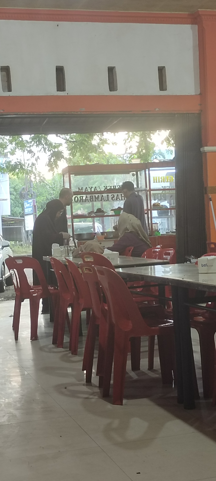 The image depicts an outdoor dining area, likely at a food stall or restaurant, in Lambaro, Indonesia. The setting appears to be illuminated by artificial lighting and natural daylight filtering through an opening above. Red plastic chairs are arranged around tables, some of which are covered with a white tablecloth and have napkins on them. In the background, a food display counter is visible, containing food items and a large pan. Three individuals are present: one person in a black hijab and dark clothing appears to be interacting with the food counter, another person in a light-colored hijab and maroon clothing is seated at a table, and a third person, a male in dark clothing, is standing and facing away from the camera near the food counter. The environment includes trees and foliage visible through the opening, suggesting an outdoor or semi-outdoor location. The overall scene suggests a casual dining establishment.