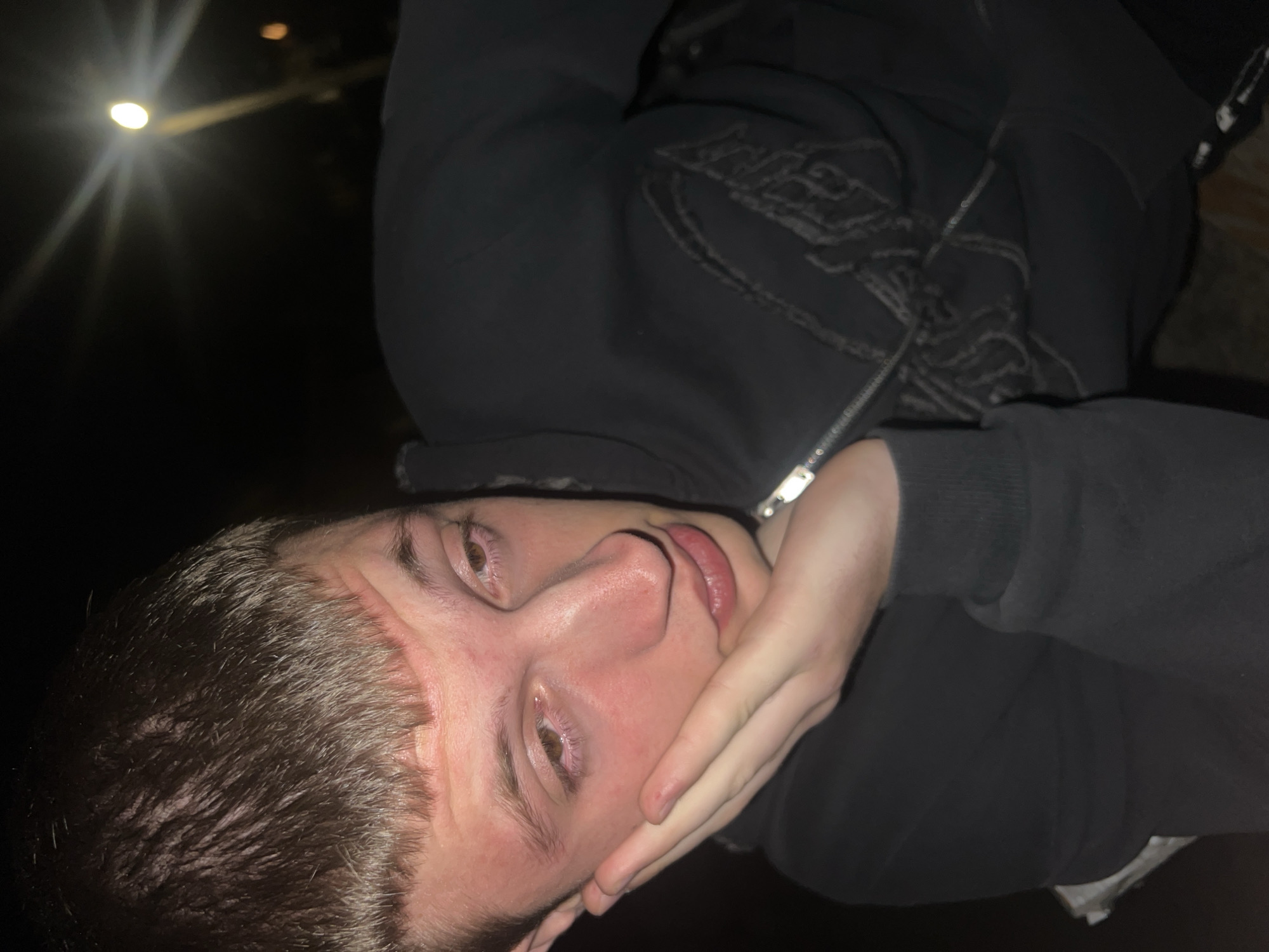 This image is a close-up, rotated portrait of a young individual, likely male, captured at night in Kochan, Bulgaria. The subject, positioned sideways, has light skin, short, dark brown hair, and appears to be looking towards the camera. He is wearing a dark-colored (likely black) hooded sweatshirt featuring a textured, indistinct graphic design on the chest and a visible metallic zipper down the front. A thin metallic chain or necklace is also visible around his neck.
His left hand is raised to his face, resting against his chin and lower cheek. His eyes appear notably red, possibly due to irritation, fatigue, or the effect of the lighting. Reddish blotches are also visible on his cheeks and forehead. His expression is somewhat neutral, leaning towards pensive or tired.
The background is almost entirely dark, save for a prominent, bright, circular artificial light source in the upper left corner, exhibiting a distinct starburst flare effect, characteristic of a street lamp or similar outdoor illumination. Another fainter, out-of-focus light source can be seen further in the background. The overall setting appears to be outdoors at night, with the artificial light providing the primary illumination for the subject. No specific text is clearly legible on the clothing.