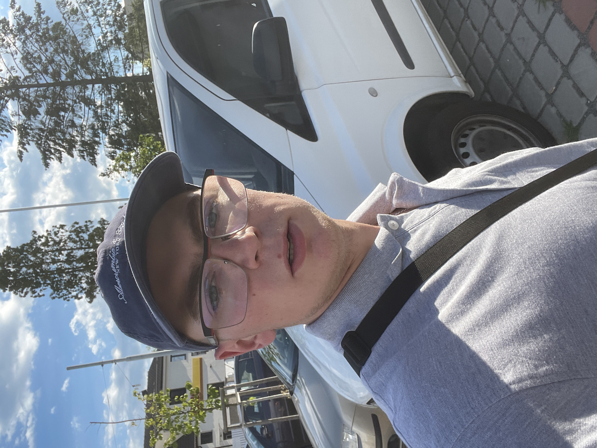 A young man wearing a grey polo shirt and a blue baseball cap with "Abercrombie New York" embroidered on it, and black-rimmed glasses, takes a selfie outdoors. He is standing in front of a white van, with a silver car visible behind him. The ground is paved with grey cobblestones. Trees and a building can be seen in the background under a blue sky with scattered clouds. The lighting suggests it is daytime.