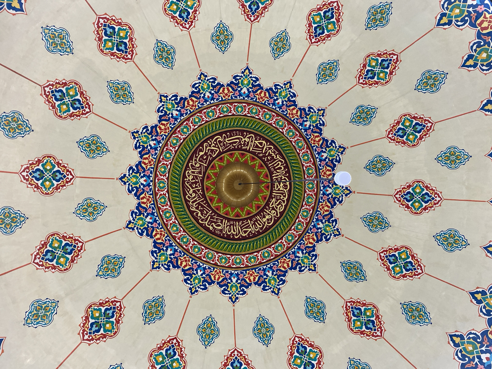 The image shows a close-up, low-angle view of the elaborately decorated ceiling of a mosque in Lhokseumawe, Indonesia. The ceiling is painted in a vibrant color palette of red, blue, green, yellow, and white, with intricate geometric patterns and Arabic calligraphy adorning its surface. A central medallion features a complex design with radiating lines, surrounding an area of calligraphy in gold. A brass-colored light fixture hangs from the center of the ceiling, with a white lightbulb visible at the bottom of the frame. The surrounding areas of the ceiling are decorated with repeating motifs of stylized floral or leaf-like shapes. The overall impression is one of rich cultural and artistic detail.