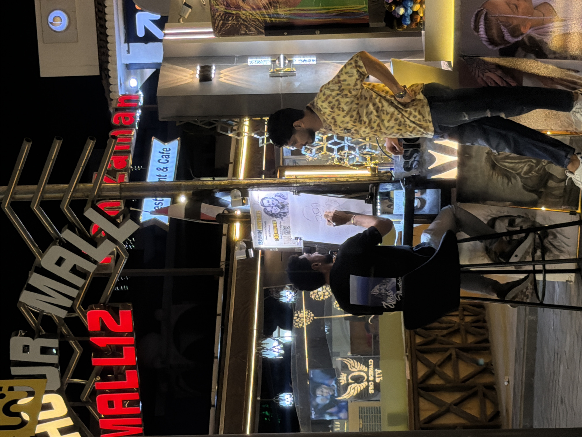 In Sharm El Sheikh, Egypt, at the "Hour Mall 12 Zaman" outdoor establishment, two men are engaged in activities related to art and commerce at night. The scene is illuminated by a combination of neon signs, string lights, and spotlights. One man, facing away from the viewer and seated on a high stool, is in the process of drawing on a large piece of paper mounted on an easel. He is wearing a black t-shirt with a mountain landscape graphic on the back and dark pants. The other man, facing left, stands beside the easel, looking towards the artist. He is wearing a yellow and black leopard-print short-sleeved shirt and dark, distressed jeans, and is resting his left hand on his hip. In the background, a lit sign reads "IBRAHIM PORTRAITS" and advertises "130 MINUTES!" for drawings. Several artworks, including portraits and a horse, are displayed around the area. Hanging lamps with intricate designs are visible above. A television screen displays an image of two people. The environment suggests a commercial street or plaza area with distinct architectural elements and decorative lighting.