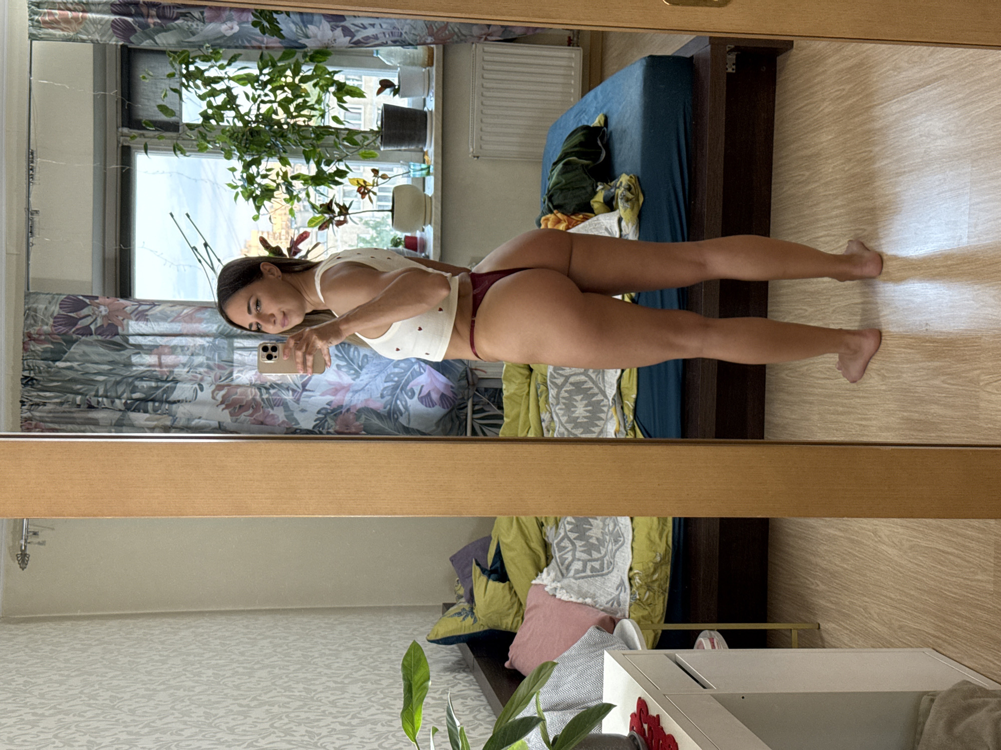 A woman is captured in a full-length mirror selfie, standing in a bedroom. She is facing away from the camera, turned to her left, and looking over her shoulder towards the mirror. She is wearing a white crop top adorned with small red hearts and a dark red thong. Her body is positioned to showcase her back and buttocks. The room's setting is visible in the reflection. A bed with unmade, brightly colored bedding dominates the background. To the left of the bed, a white dresser is partially visible. A window with floral curtains is behind the bed, with several potted plants on the windowsill. The flooring is light-colored wood. The overall impression is that of a casual, personal moment captured in a domestic space.