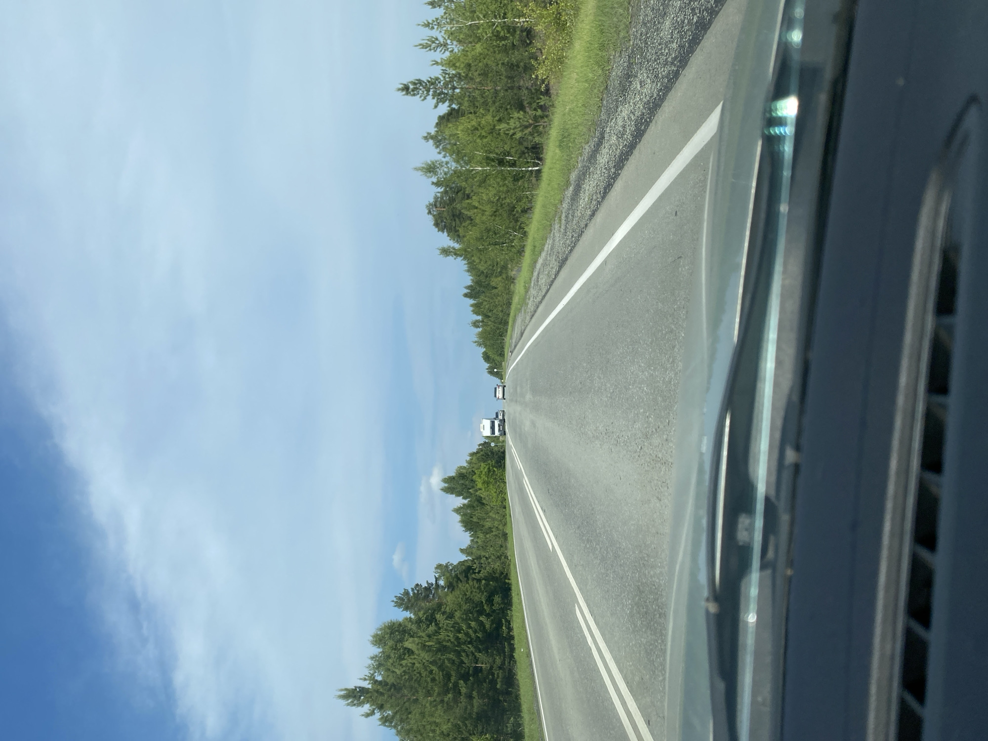 This image captures a view from inside a vehicle traveling on a paved highway. The road is marked with white lines and appears to be a two-lane road with traffic moving in the same direction. On either side of the road, there is a dense forest with a mix of coniferous and deciduous trees. The sky is partly cloudy with a blue sky visible. In the distance, several vehicles are visible on the highway, including a white truck and a couple of cars. The foreground shows the dashboard and windshield of the vehicle, with some reflections on the glass. The scene suggests a drive through a rural or forested area, possibly on a highway in Zilair, Russia, given the context provided. The overall impression is one of travel through a natural landscape under a daytime sky.