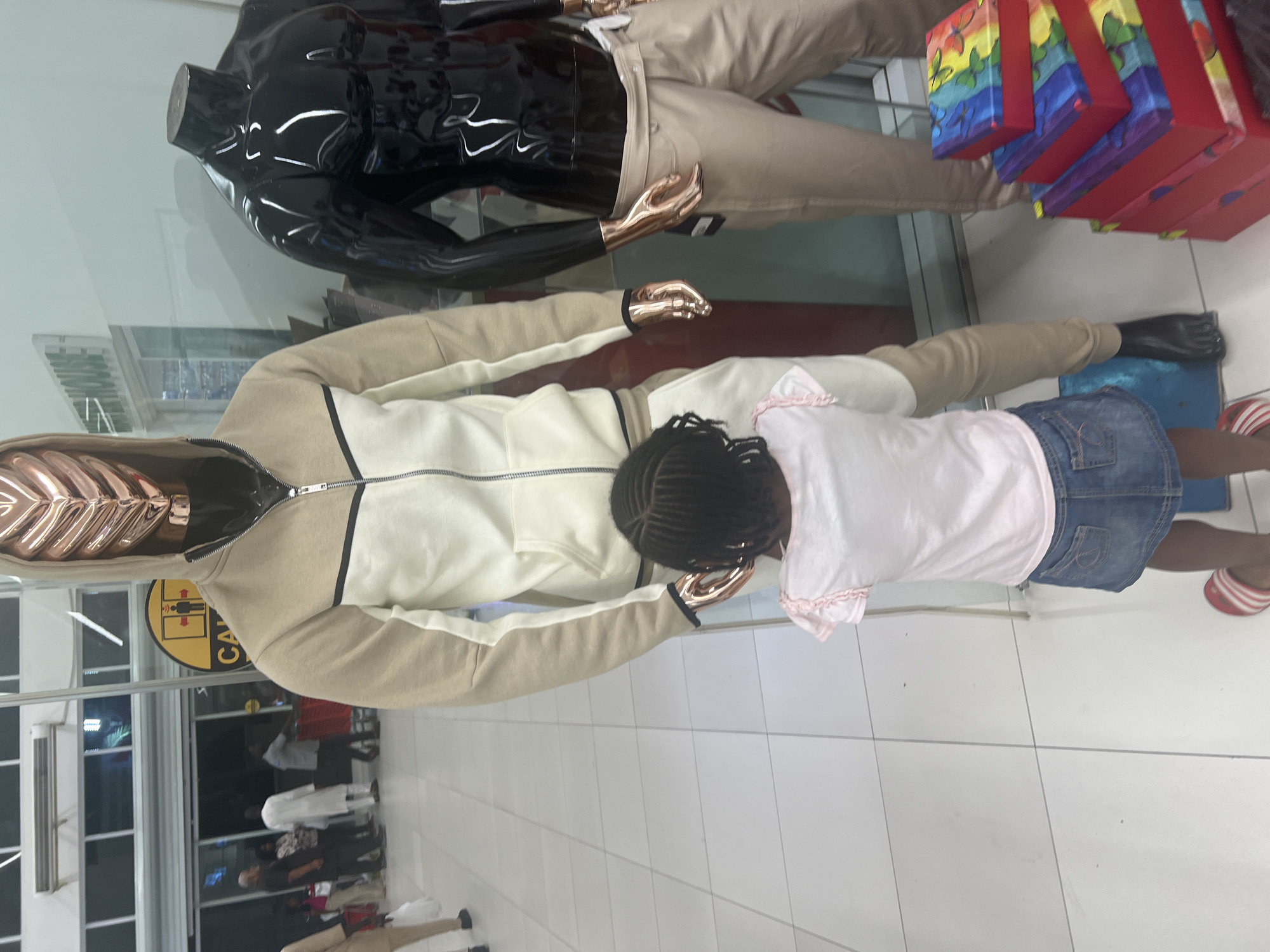 A child with braided hair stands in front of a mannequin dressed in a beige and white hooded jacket with a zipper. The child wears a light pink t-shirt and a denim skirt. Mannequins are displayed in a storefront window behind them, including a shiny black torso and a mannequin dressed in khaki pants and a dark jacket, standing amongst stacked, colorful boxes. The flooring is white tile. The environment appears to be inside a clothing store, and outside, one can see reflections and other shoppers. While the city of Abuja, Nigeria, is not discernible from the image itself, the prompt mentions that it is the location.